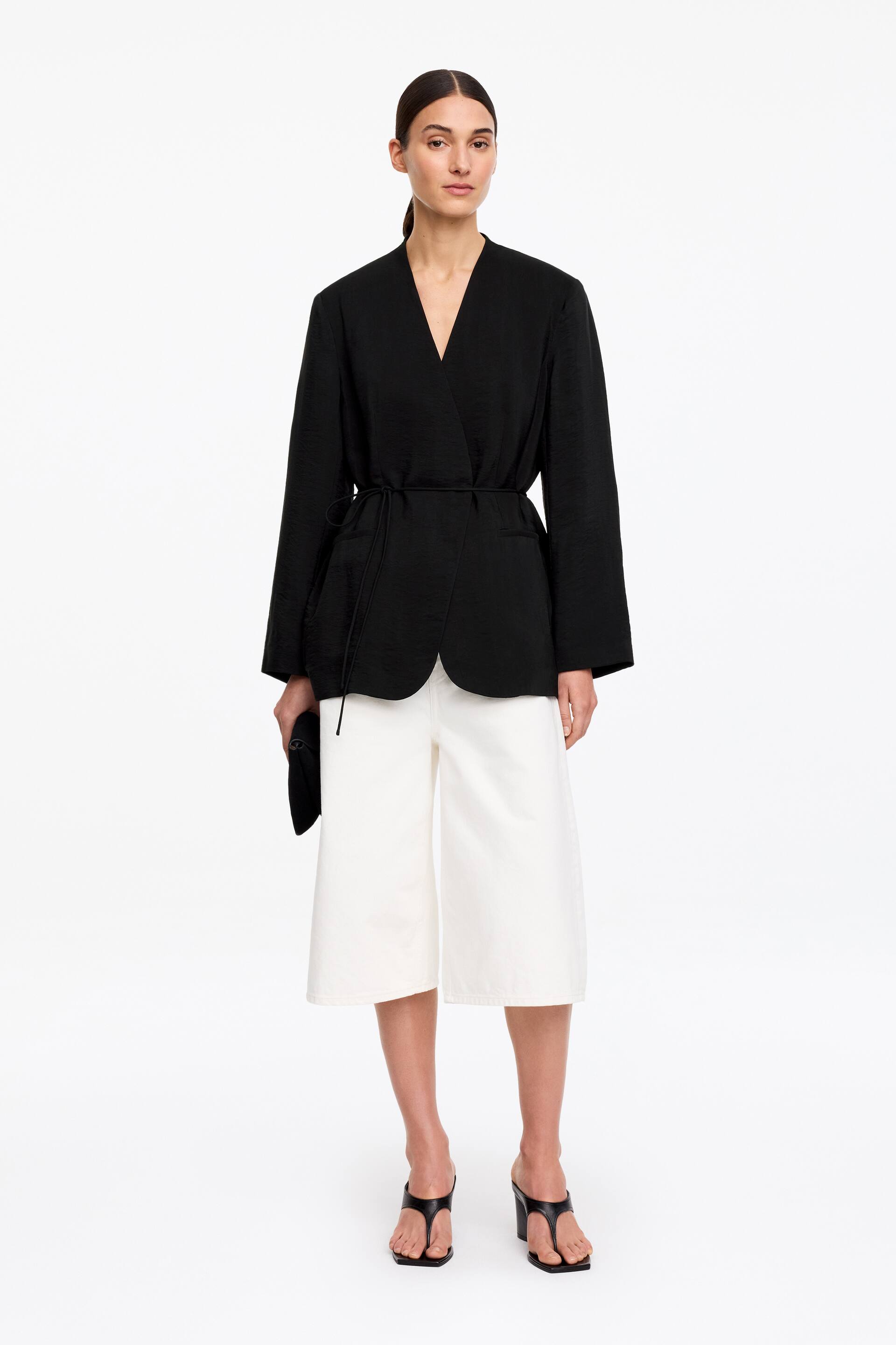 Zobrazit větší obrázek: Modelka má na sobě černé zavinovací sako, které je charakteristické výstřihem do V, páskem na zavázání vpředu a lištovými kapsami. Modelka také drží černou psaníčko vedle saka.