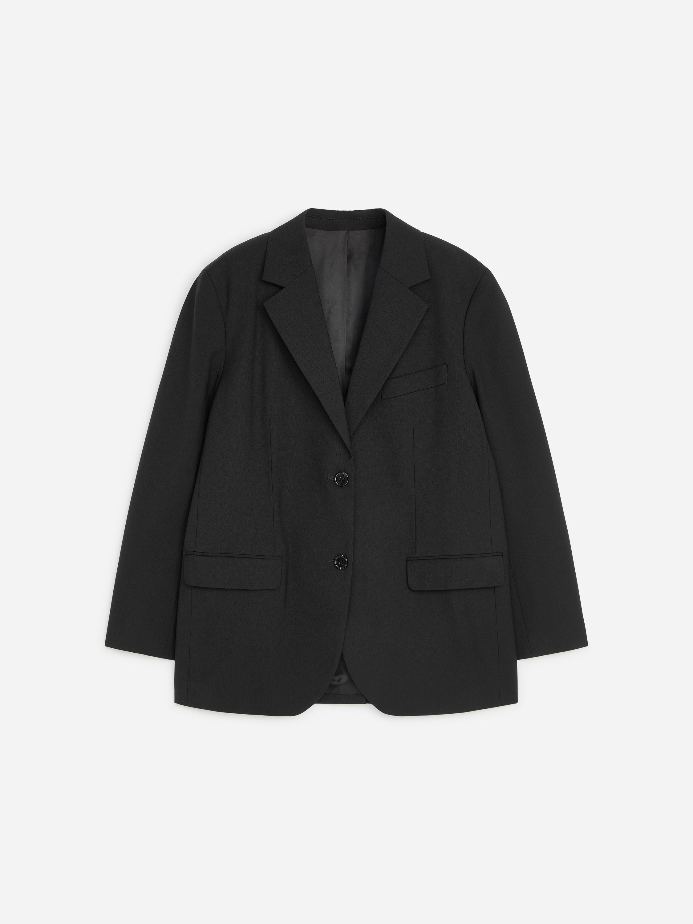 La modella indossa un blazer sartoriale nero dalla vestibilità rilassata, abbinato a una T-shirt bianca e a jeans scuri a gamba ampia. Il blazer monopetto presenta rever classici, chiusura frontale con due bottoni e tasche con patta, mentre la modella tiene in una mano una piccola clutch nera.