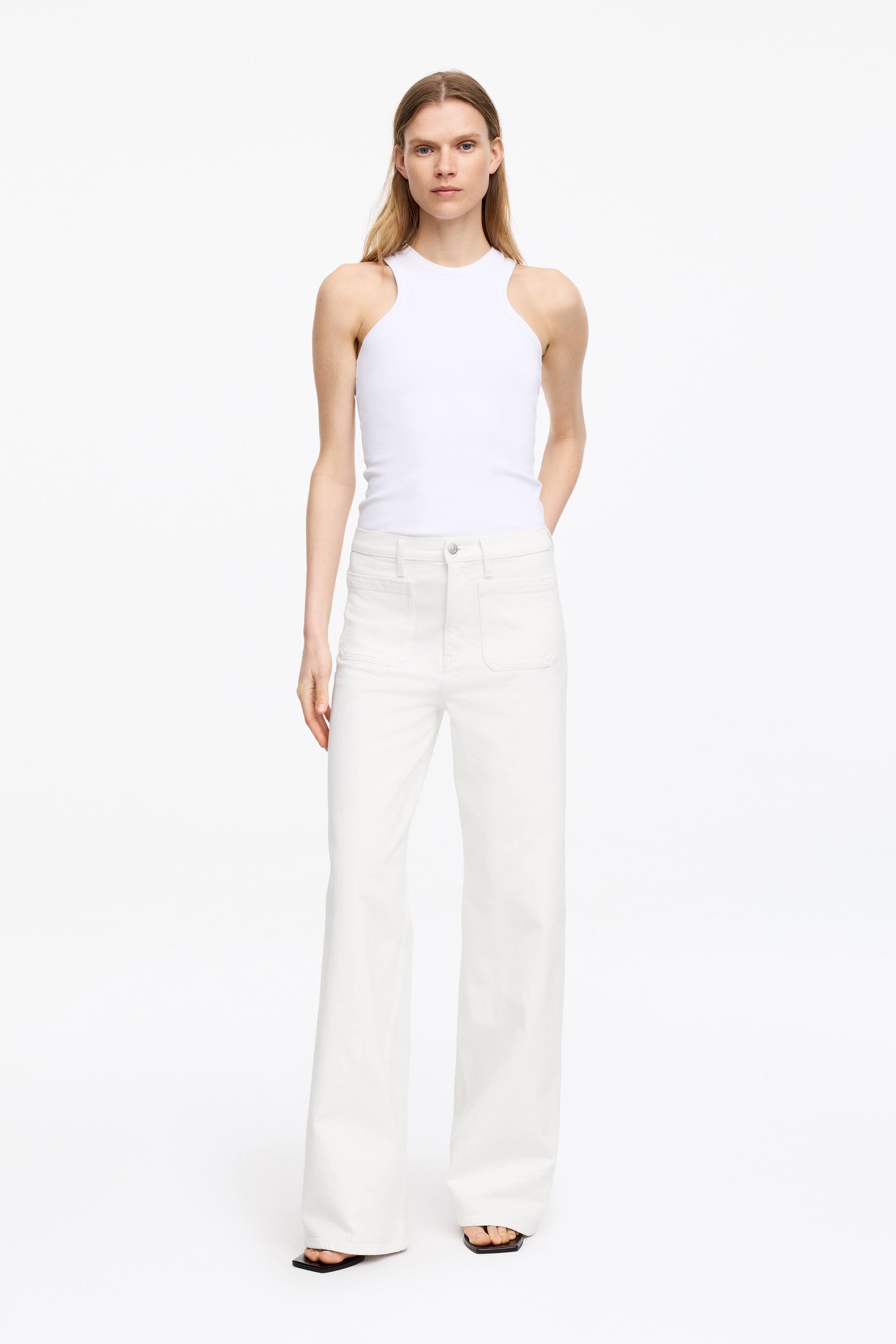 Visualizza immagine più grande: I jeans LUPINE sono bianchi, a vita alta, con una vestibilità aderente e gambe svasate. Le tasche frontali applicate sono visibili. La modella indossa i jeans.