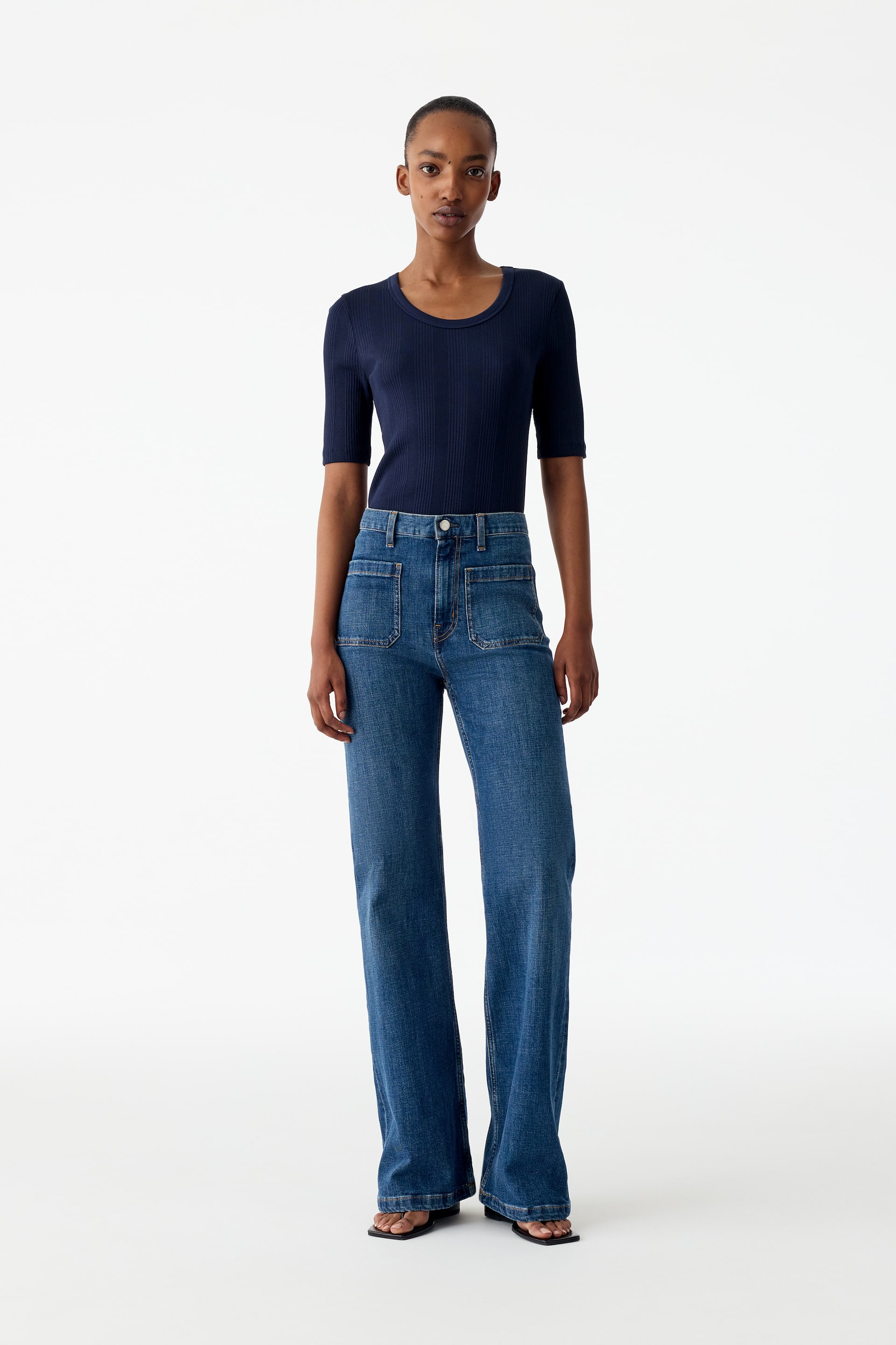 Visualizza immagine più grande: La modella indossa jeans in denim blu con vita alta, tasche applicate sul davanti e gamba svasata. I jeans sono abbinati a una maglietta a costine blu scuro a maniche corte, infilata داخل.