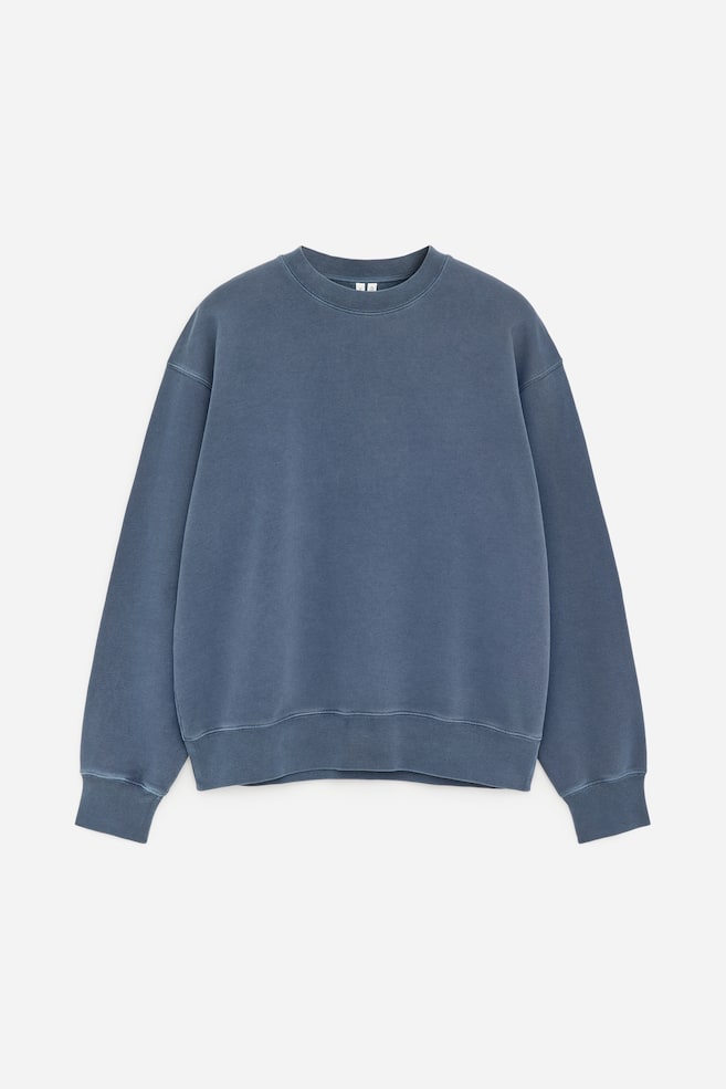 El frente de una sudadera azul grisáceo muestra un corte holgado con hombros ligeramente caídos. Presenta un aspecto desteñido y teñido en prenda, junto con un cuello redondo, puños acanalados y un dobladillo acanalado. - 1