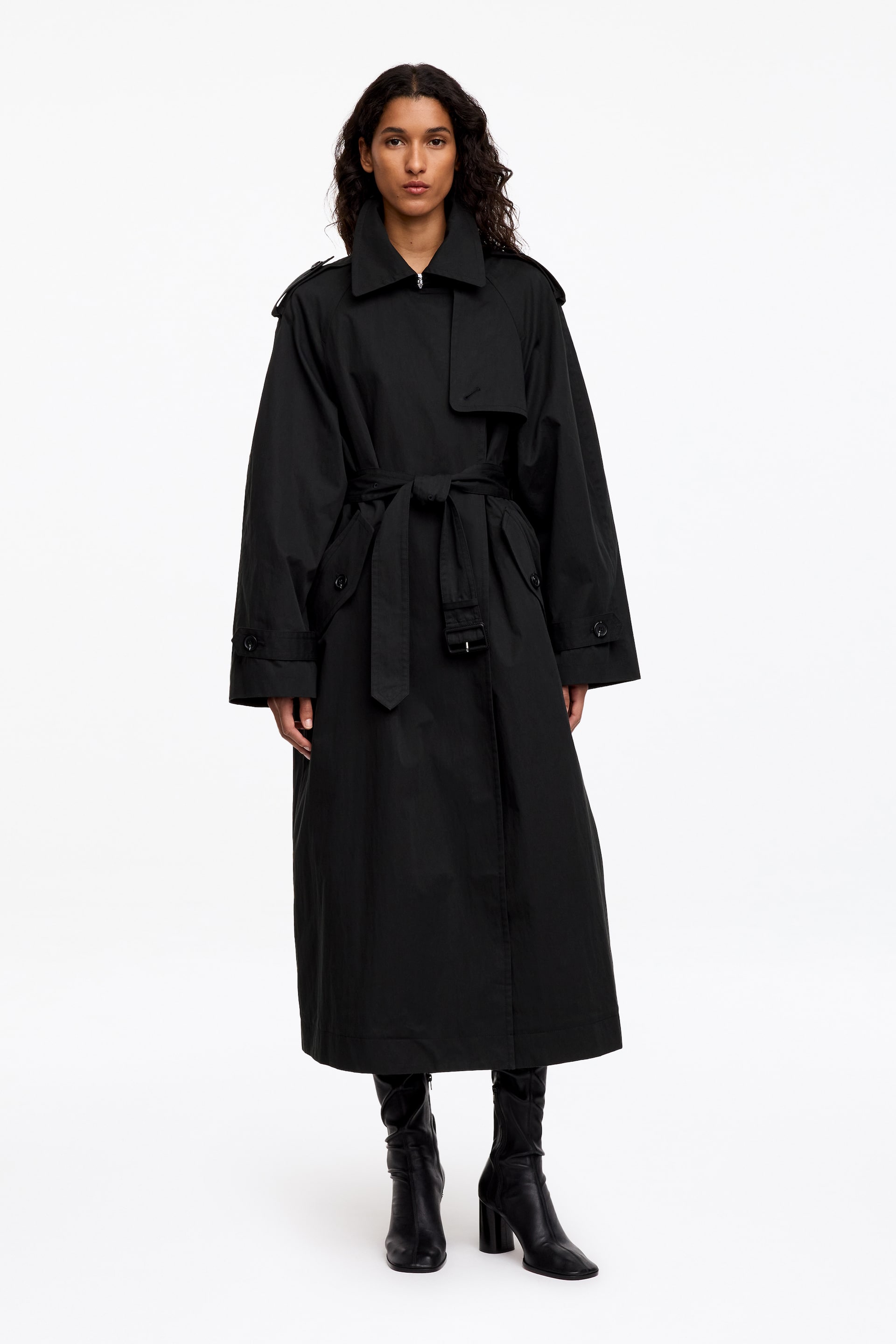 Visa större bild: En svart, oversized trenchcoat bärs av modellen och har en bred spetsig krage, axelklaffar, en vindslå och justerbara ärmuppslag. Kappan knyts i midjan med ett matchande tygbälte och kombineras med svarta stövlar med klack.