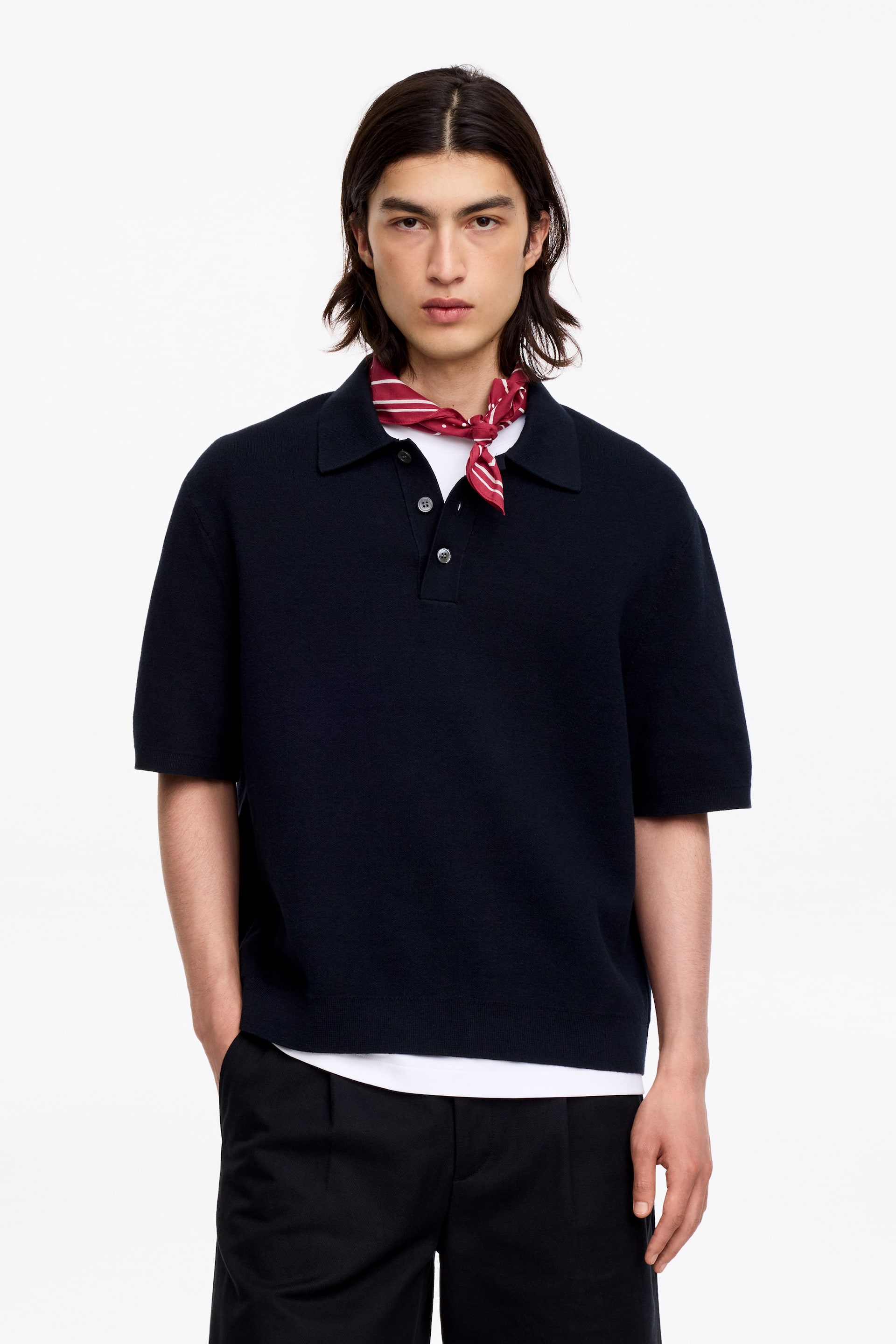 Se større billede: En marineblå strikket poloshirt vises forfra, med en spids krave, en tre-knaps stolpe, korte ærmer og en lige kant. En hvid t-shirt er lagdelt nedenunder, og et rød- og hvidstribet bandana er bundet om halsen.