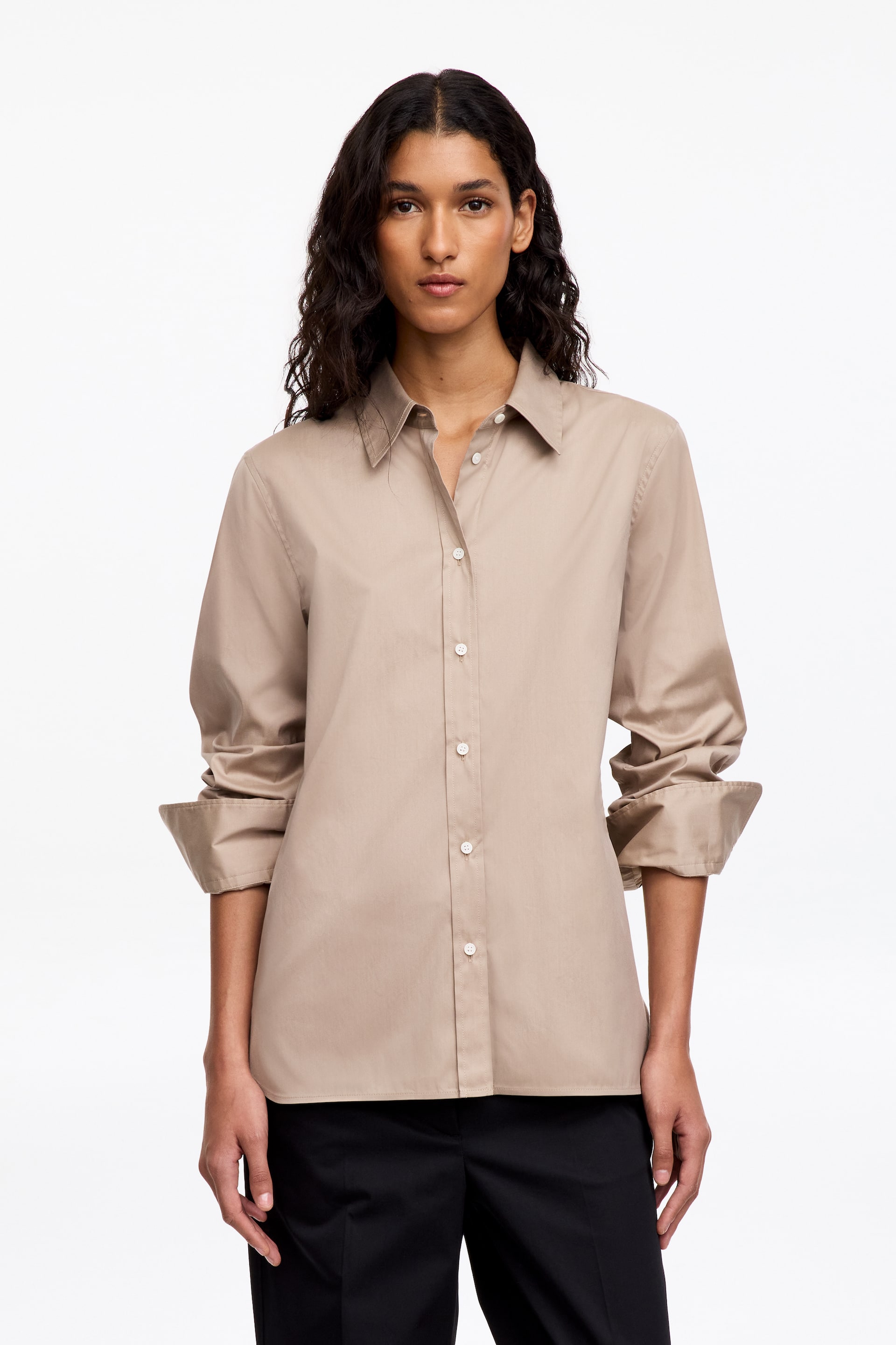 Agrandir l'image: Une chemise en coton beige, coupe slim, est portée par le mannequin, avec un col classique, une fermeture boutonnée sur le devant et des manches longues aux poignets retroussés. La chemise est portée non rentrée et associée à un pantalon noir.