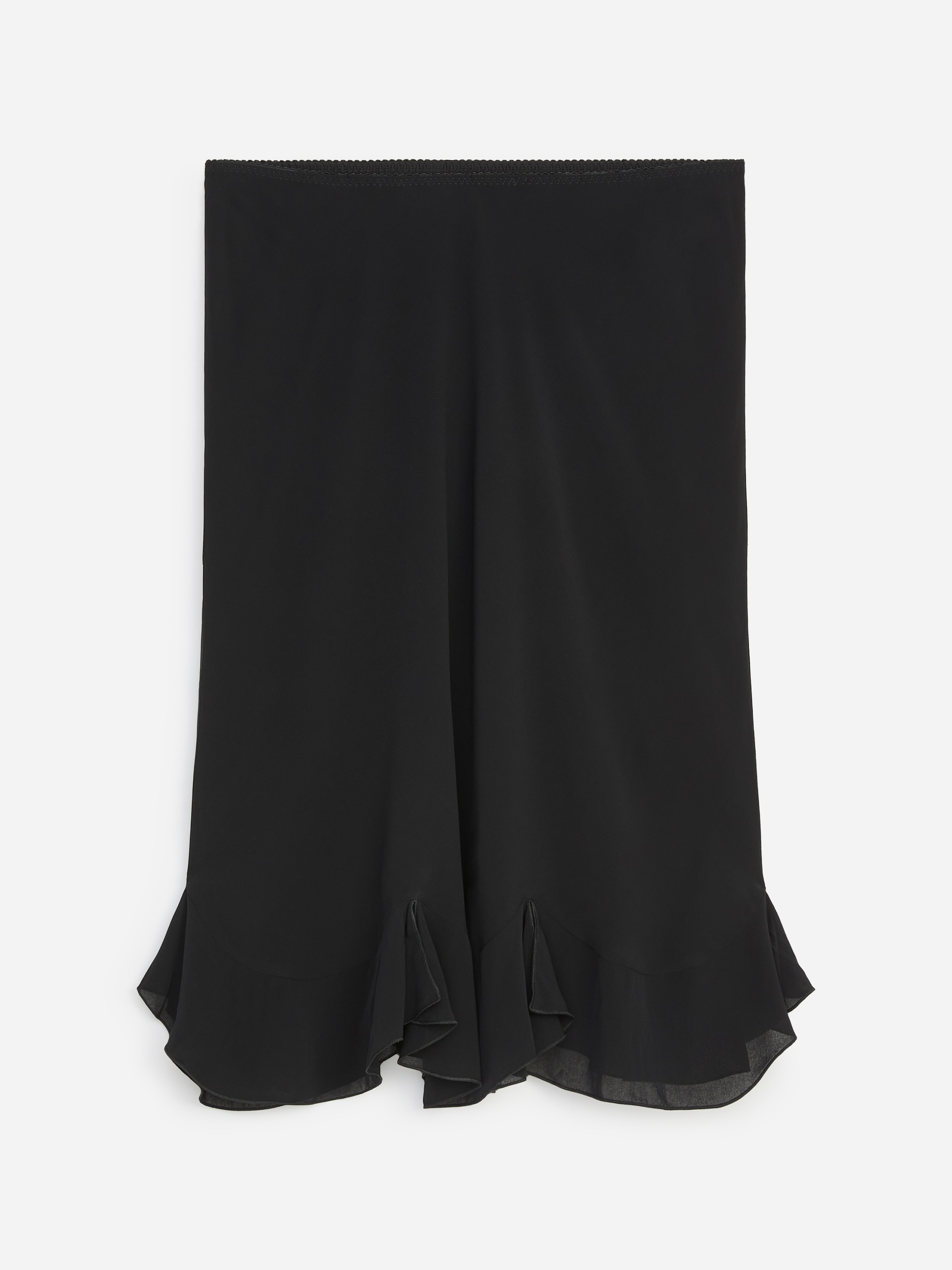 La modelo lleva una falda midi negra, con una silueta de corte al bies suavemente drapeada y un bajo de volante calado con acabado festoneado. La falda se combina con un top blanco de manga larga holgado, parcialmente metido por dentro de la cintura, y se completa con botas altas de cuero negro.