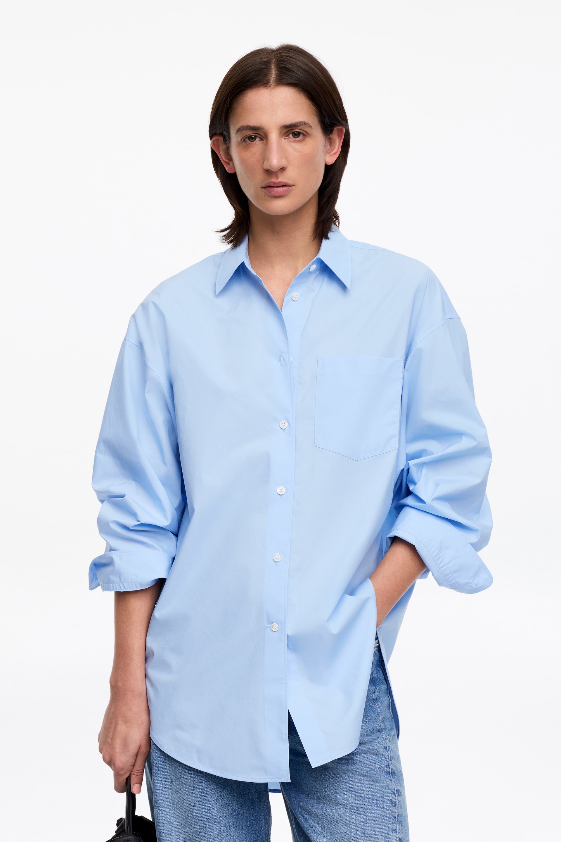 Agrandir l'image: Une chemise en coton bleu clair surdimensionnée, avec un col, une fermeture complète à boutons sur le devant et une poche poitrine unique, est portée par le mannequin. Ses manches surdimensionnées sont retroussées, et le mannequin place une main dans la poche de la chemise.