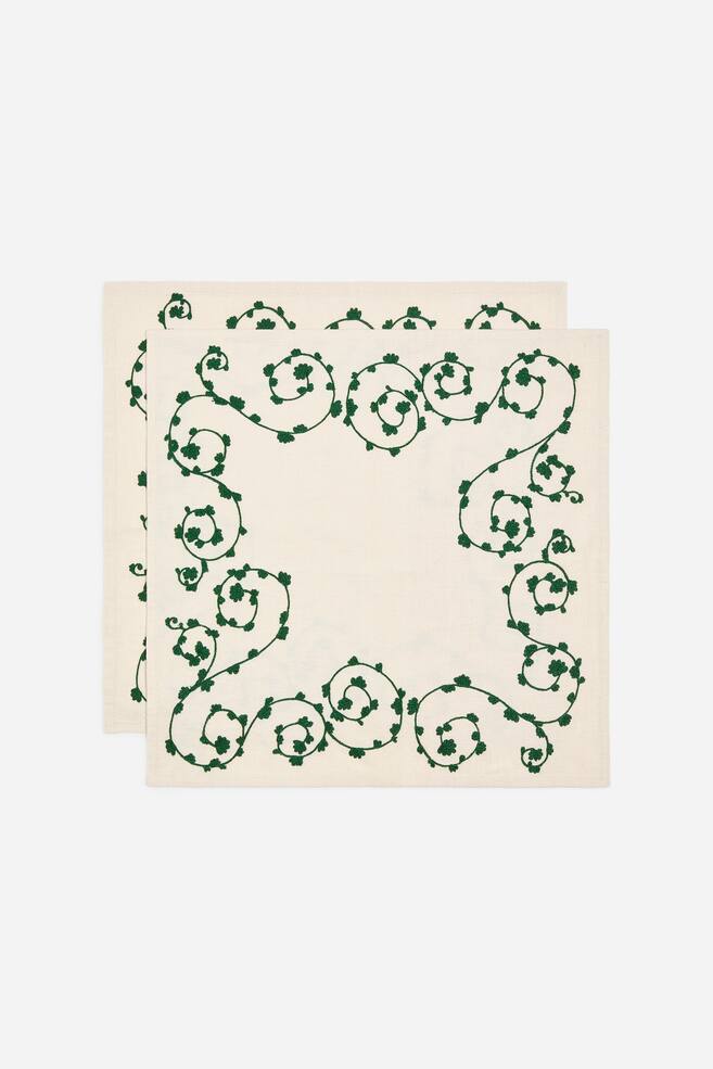 TvÃ¥ fyrkantiga servetter i benvitt tyg visas plant frÃ¥n en frontvÃ¤nd vinkel, staplade nÃ¥got fÃ¶rskjutet ovanpÃ¥ varandra. Varje servett har en grÃ¶n broderad bÃ¥rd med slingrande vinrankor och smÃ¥ blad som ramar in en enfÃ¤rgad mitt. - 1