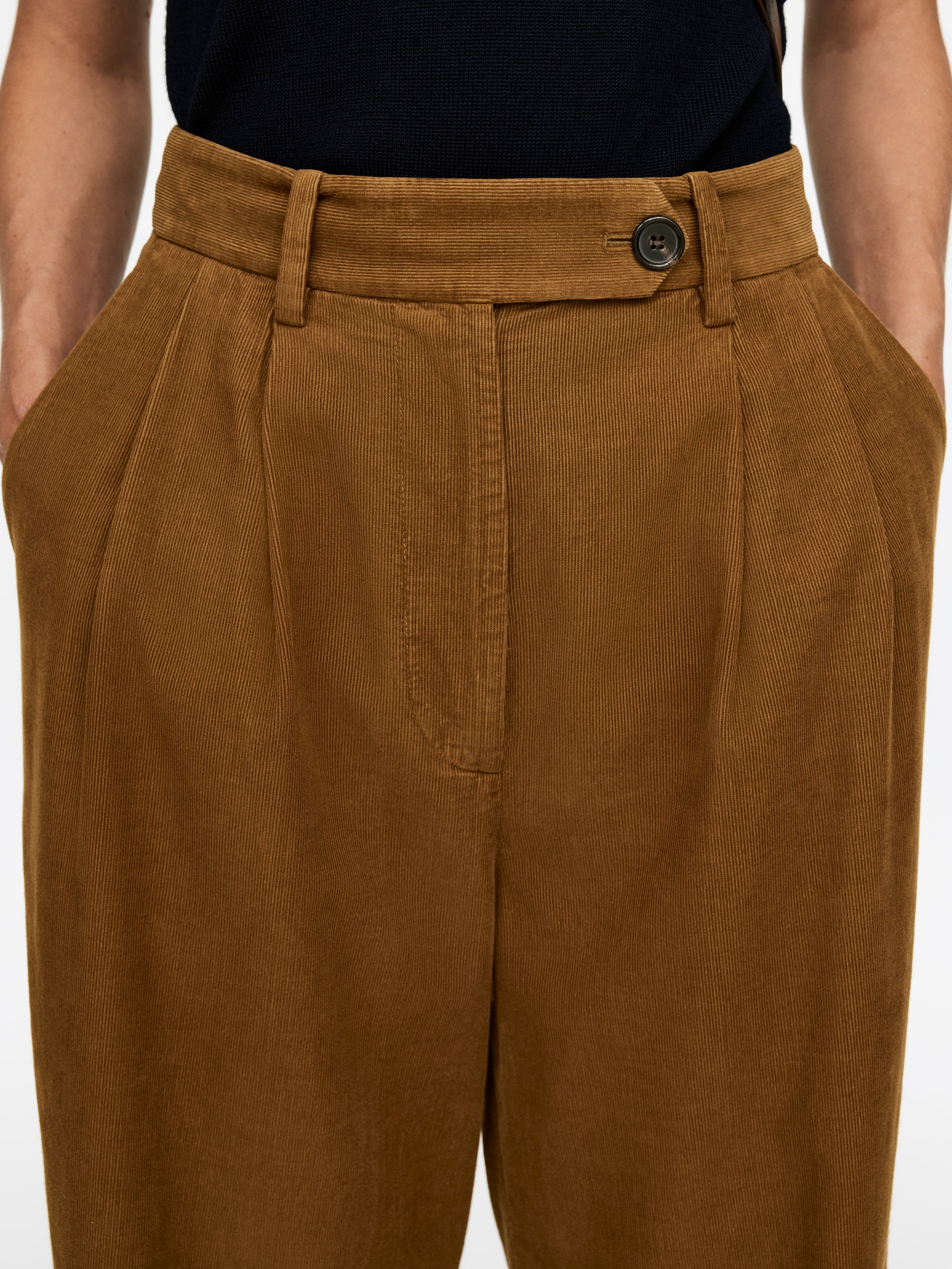 View larger image: Brown corduroy pants are featuring a wide waistband with belt loops and a single dark button closure on an extended tab. Two deep pleats are detailing the front, and side pockets are visible.