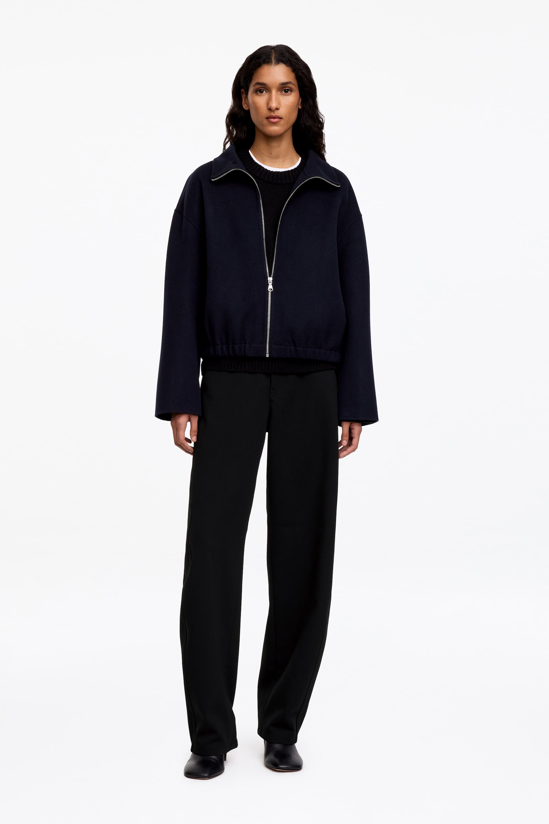 Visa större bild: Dessa svarta ullbyxor, med en avslappnad, mjukt böjd barrel-leg-silhuett, bärs av modellen. Byxorna kombineras med en marinblå zip-up-jacka, en svart crewneck-tröja och svarta läderskor.