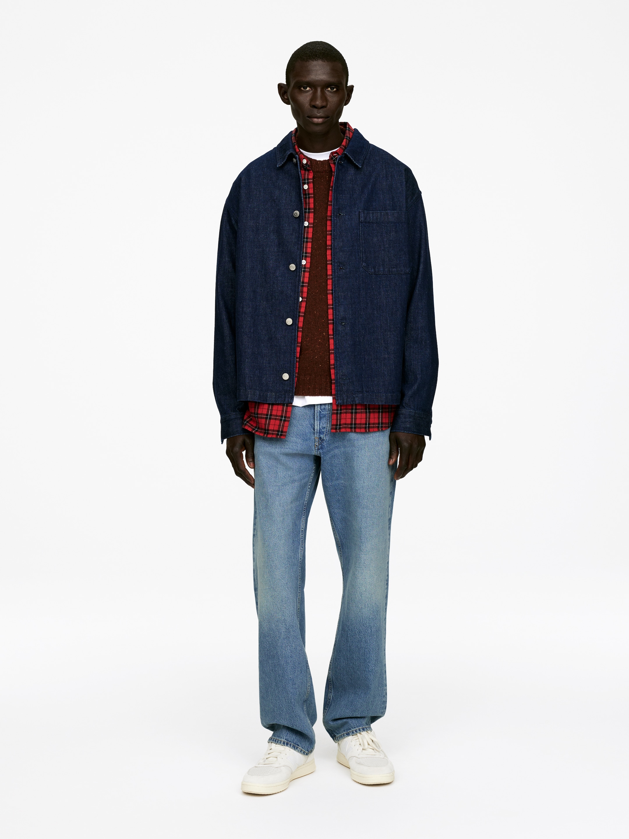Grotere afbeelding bekijken: Een casual gelaagde outfit wordt gepresenteerd in een vooraanzicht, met een donkerblauwe denim overshirt met een knoopsluiting aan de voorkant en een enkele borstzak, gedragen open over een bruine gebreide trui met ronde hals en een rood en zwart geruit overhemd met knopen. Licht gewassen jeans met een relaxte pasvorm worden gecombineerd met witte en beige vetersneakers, waarmee het ensemble wordt voltooid.