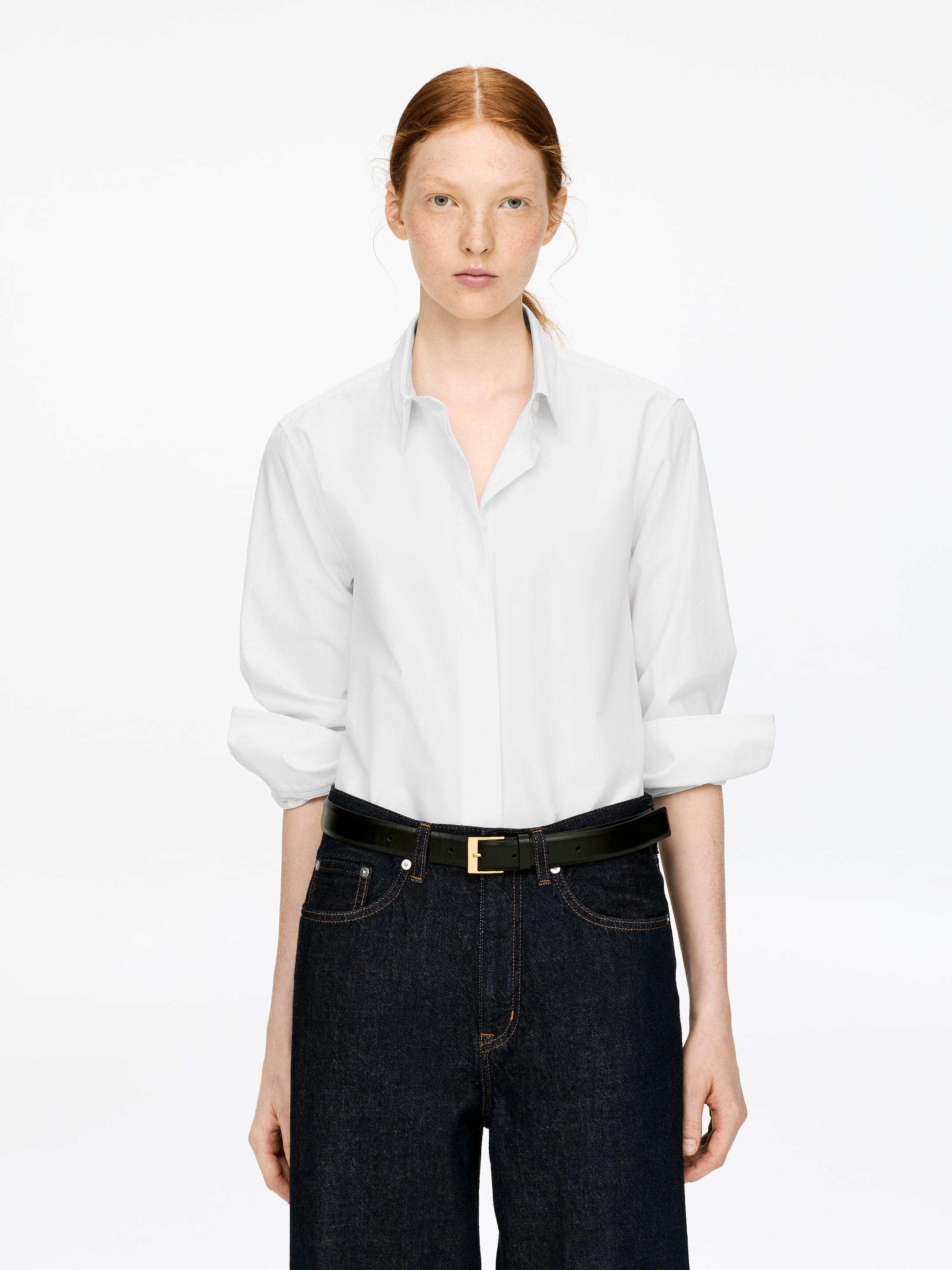Visualizza immagine più grande: Una classica camicia bianca in cotone con una vestibilità regolare e una chiusura con bottoni nascosta è indossata dalla modella. Le maniche lunghe della camicia sono mostrate con i polsini risvoltati e arrotolati, e la camicia è infilata in pantaloni di denim scuro, che una cintura nera fissa in vita.