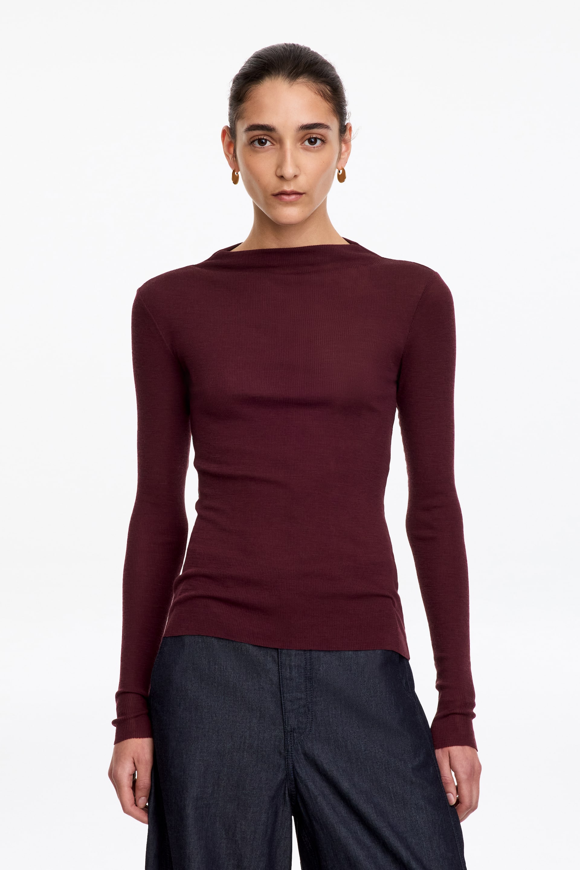 Visa större bild: En vinröd, fint ribbstickad topp med hög båtringning och långa ärmar bärs av modellen. Den figursydda toppen är stylad till mörka denimbyxor.
