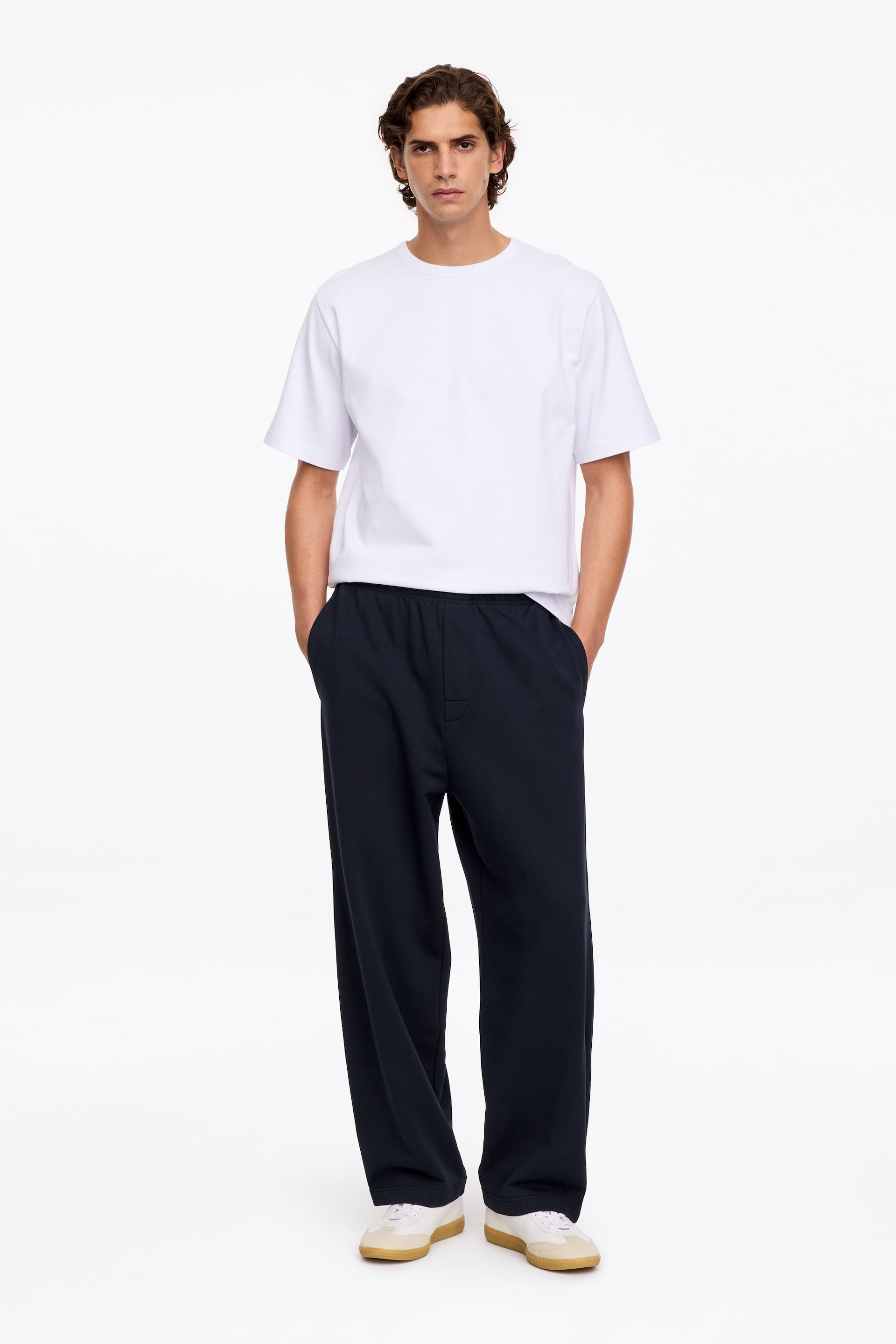Преглед на по-голямо изображение: Тъмносини трикотажни панталони с relaxed fit, с еластичен колан и имитация на цип, са облечени от модела, който е в изправена поза с ръце в страничните джобове. Панталоните са комбинирани с обикновена бяла тениска с обло деколте и бели маратонки.