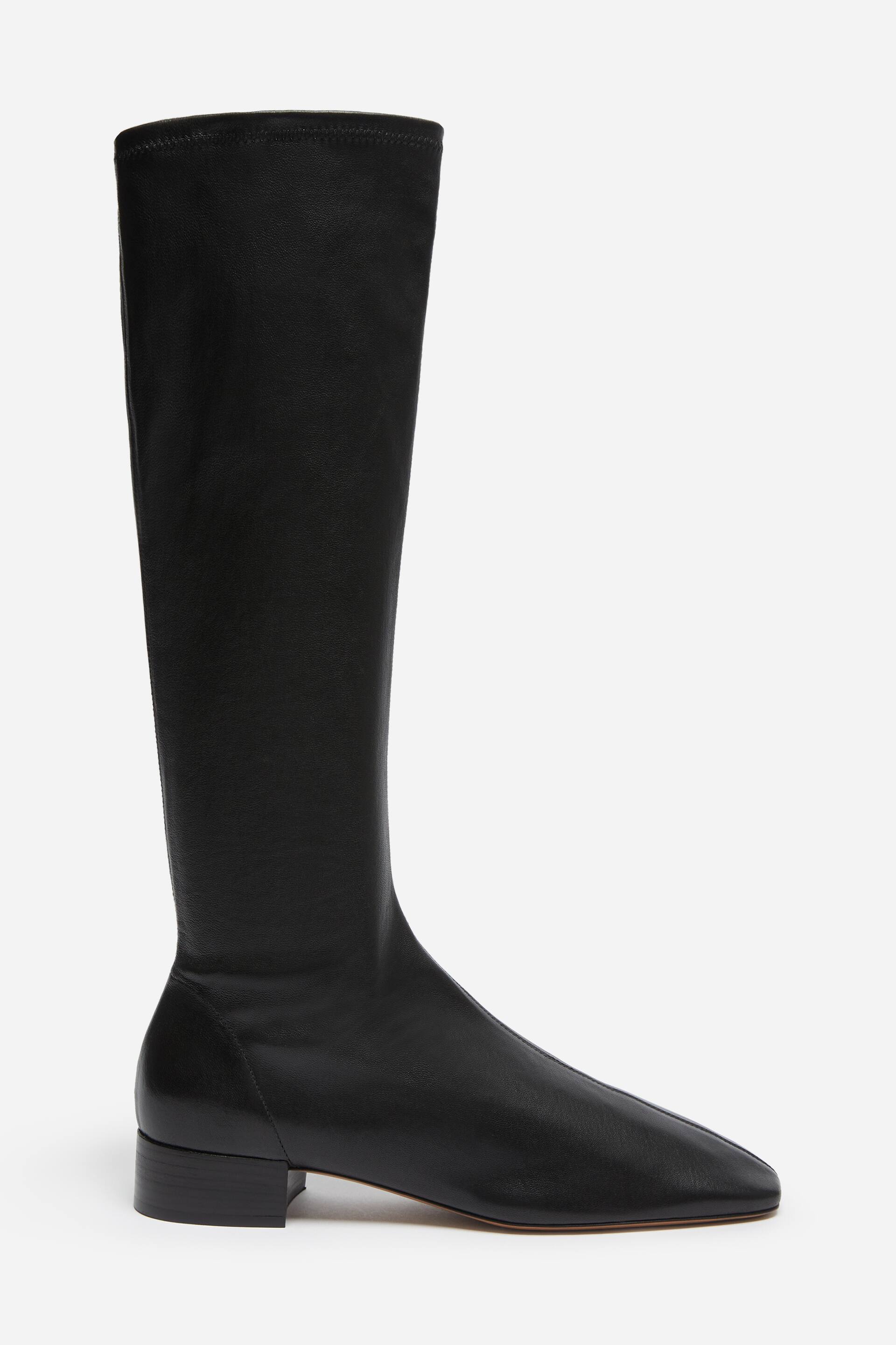 Größeres Bild ansehen: Ein schwarzer kniehoher Lederstiefel ist in einer seitlichen Profilansicht nach rechts ausgerichtet zu sehen. Er verfügt über einen weichen, körpernah anliegenden Schaft und ein Design mit eckiger Spitze und Mittelnaht. Der Stiefel ruht auf einem niedrigen Blockabsatz und einer schmalen Sohle, wodurch seine klare, minimalistische Silhouette betont wird.
