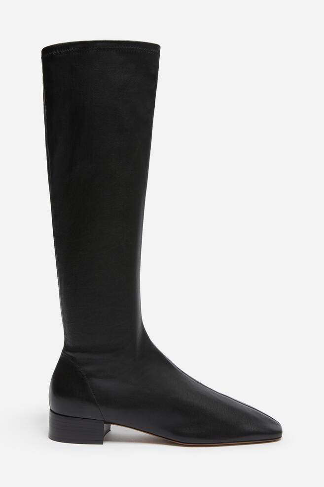 A black leather knee-high boot is shown in side profile facing right, featuring a soft, form-fitting shaft and a square-toe design with a central seam. The boot rests on a low block heel and a slim sole, highlighting its clean, minimalist silhouette. - 1