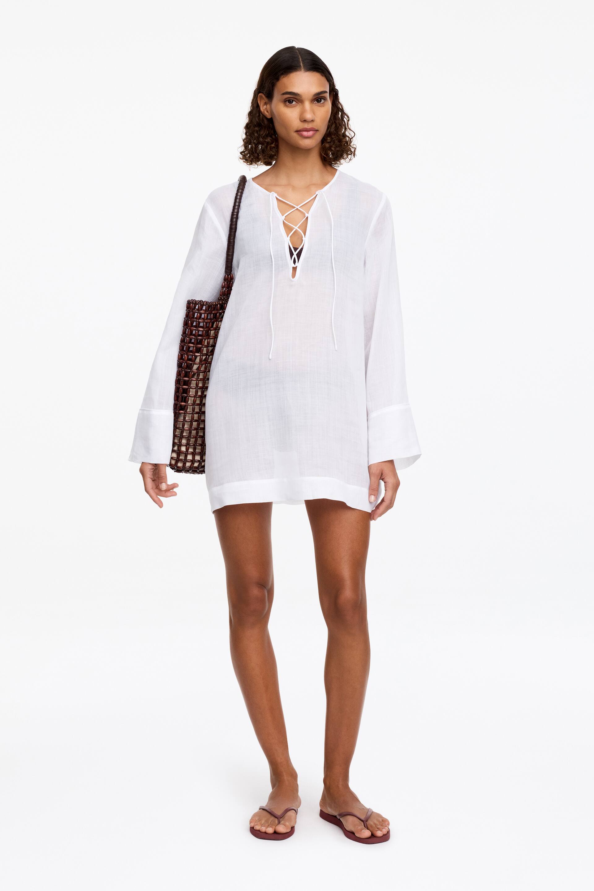 Ver imagen más grande: La modelo lleva un vestido blanco de playa como cubrebañador, que presenta una silueta relajada, mangas largas y un cierre de cordones en el escote en V del frente, con un bikini negro visible debajo. La modelo también lleva un bolso de mano tejido y unas chanclas burdeos.