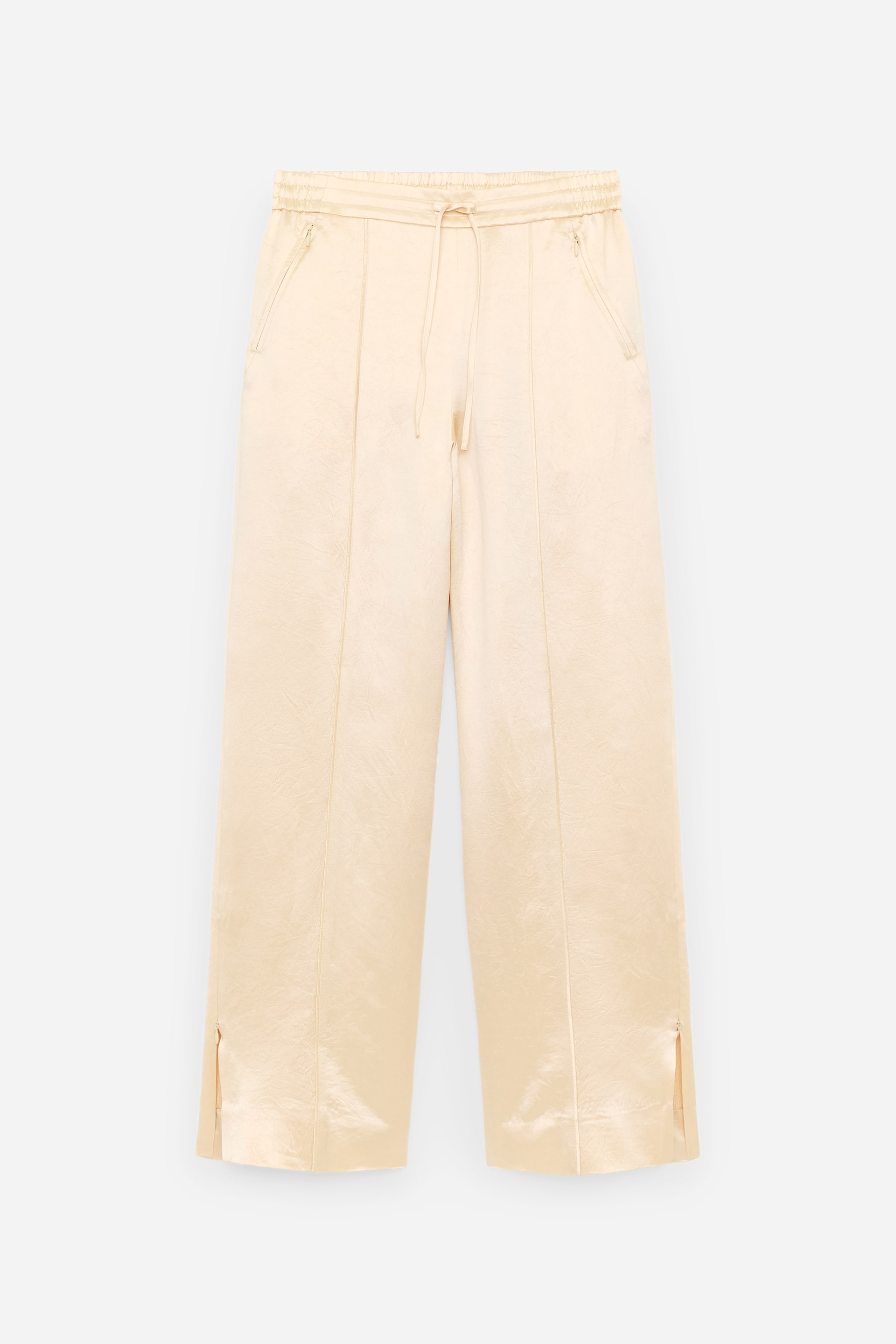 Agrandir l'image: Les pantalons crème de face sont confectionnés dans un tissu lustré subtilement froissé, dotés d'une taille élastiquée avec cordon de serrage et de deux poches avant inclinées. Des coutures verticales parcourent le centre de chaque jambe large, se terminant par de petites fentes latérales à l'ourlet.