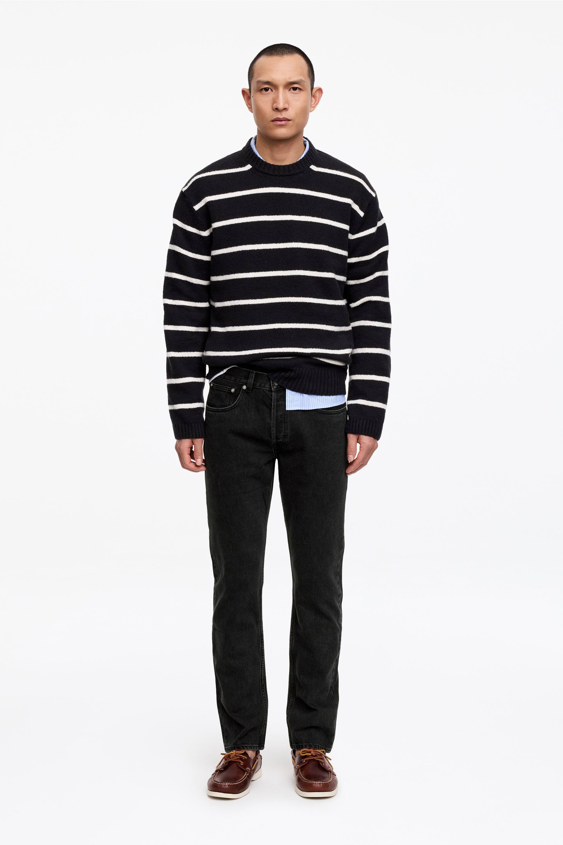 Agrandir l'image: Un pull à col rond rayé noir et blanc, avec de larges rayures horizontales, une coupe décontractée, et des poignets et un ourlet côtelés, est porté sur une chemise à col rayé bleu clair et blanc. Un jean droit gris foncé est porté, accompagné de chaussures bateau en cuir marron avec des semelles blanches et des lacets marron.