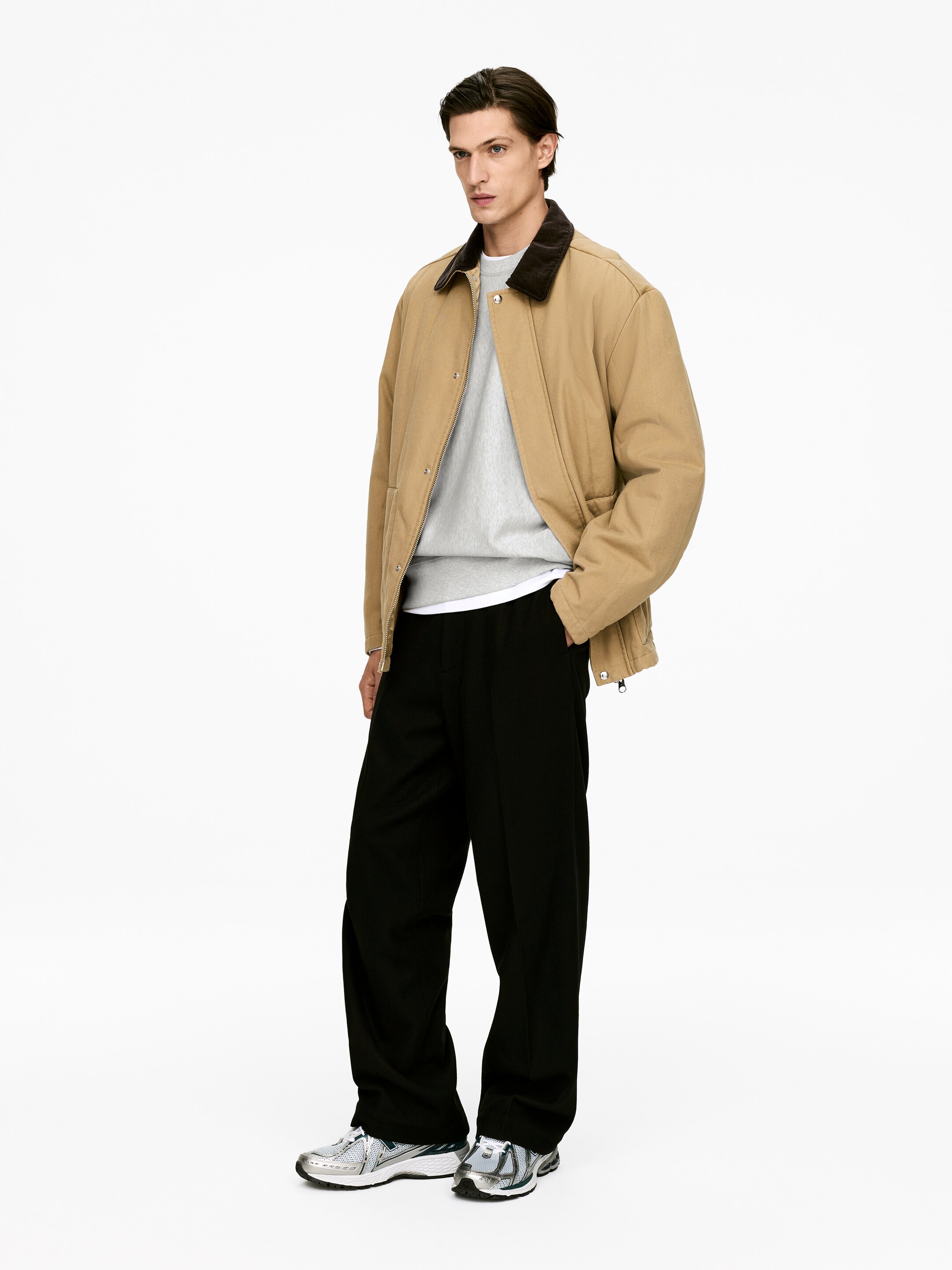 Ingrandisci l'immagine: Una giacca stile workwear color beige, con un colletto in velluto a coste marrone scuro e chiusura frontale con bottoni automatici, è presentata da un angolo leggermente obliquo, sovrapposta a una felpa girocollo di colore grigio chiaro e una t-shirt bianca. Pantaloni neri a gamba larga sono abbinati a sneakers sportive di colore argento e grigio.