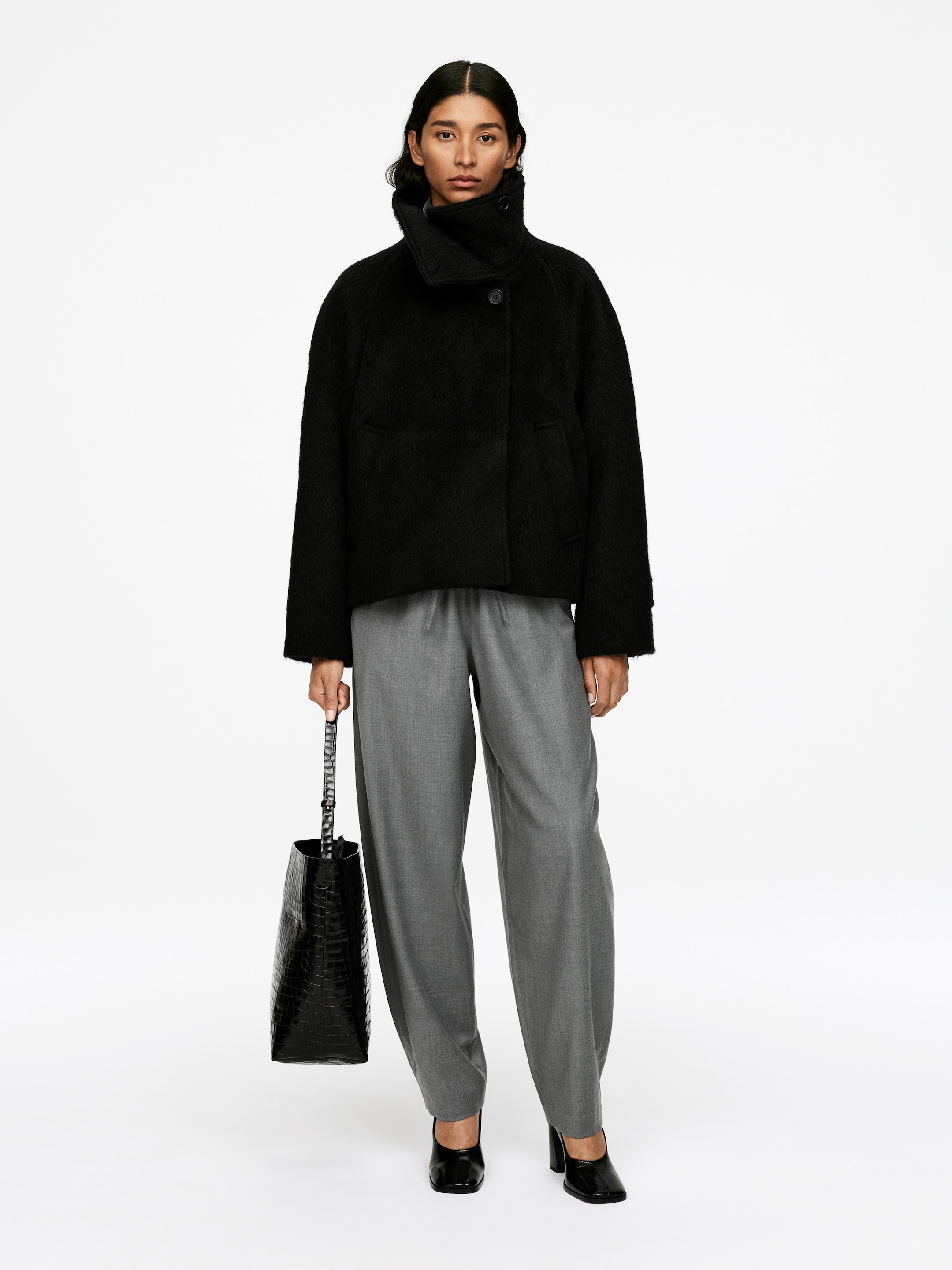 Agrandir l'image: Une veste noire, texturée, courte avec un col montant, boutonnée au cou, et des manches larges est portée avec un pantalon gris à jambes larges et coupe décontractée, doté d'une taille à cordon de serrage visible. La tenue est complétée par des chaussures noires à bout carré et talons blocs, ainsi qu'un sac seau noir embossé façon crocodile.