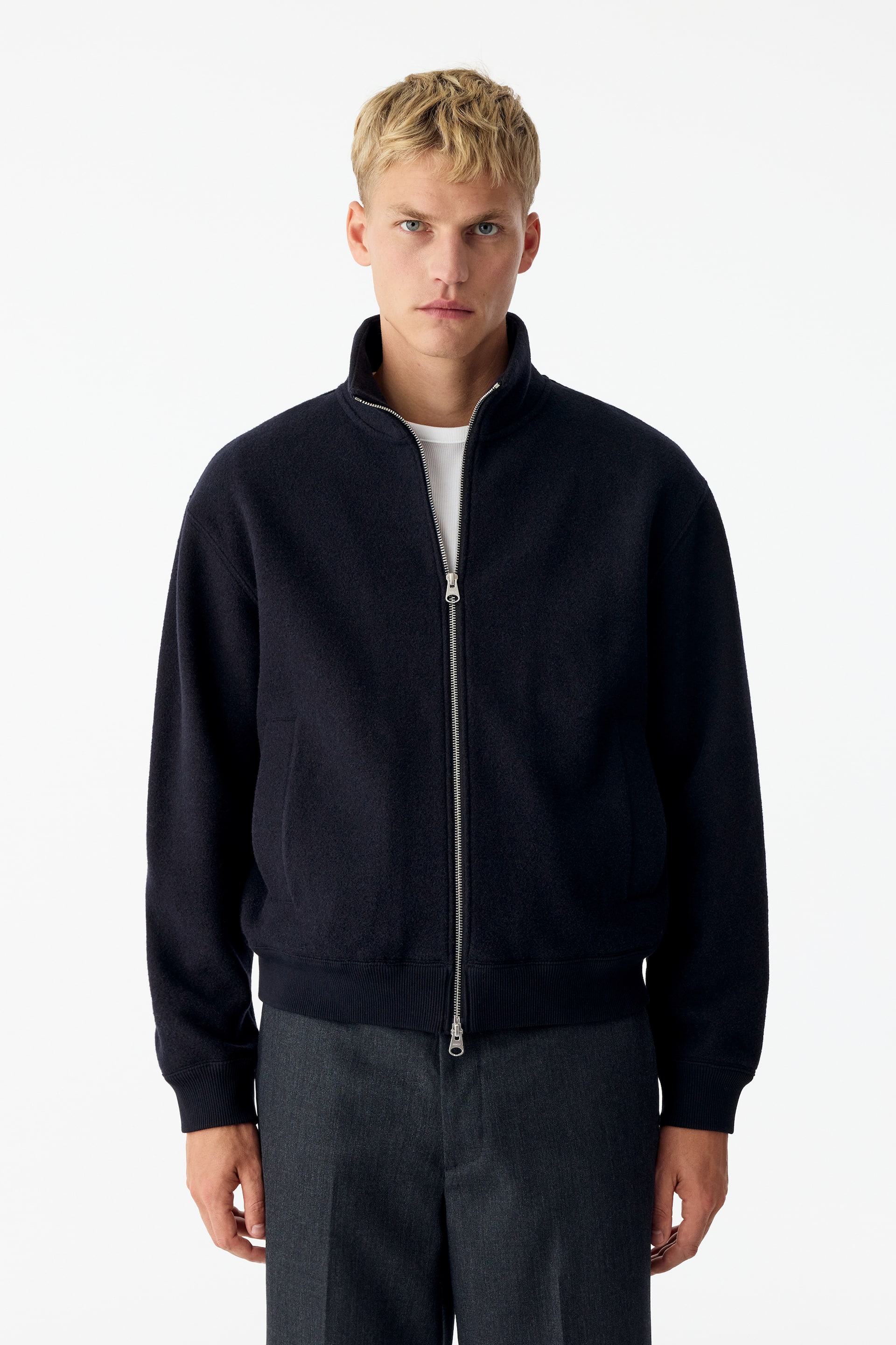 View larger image: A dark navy wool-blend jacket featuring a high collar, a silver two-way zip fastening, and ribbed trims is being worn by the model. The jacket is layered over a white T-shirt and paired with dark grey trousers.