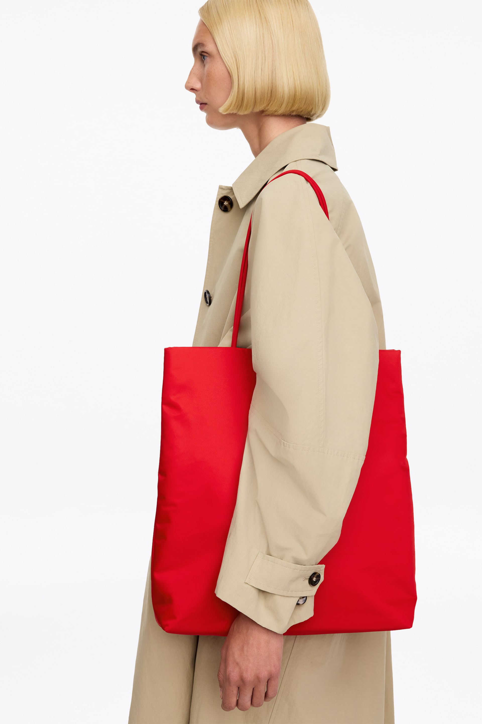 View larger image: A bright red, padded nylon tote bag with thin, rounded shoulder straps is being carried over the model’s shoulder. The model is wearing a light beige trench coat, with the rectangular bag positioned under the arm.
