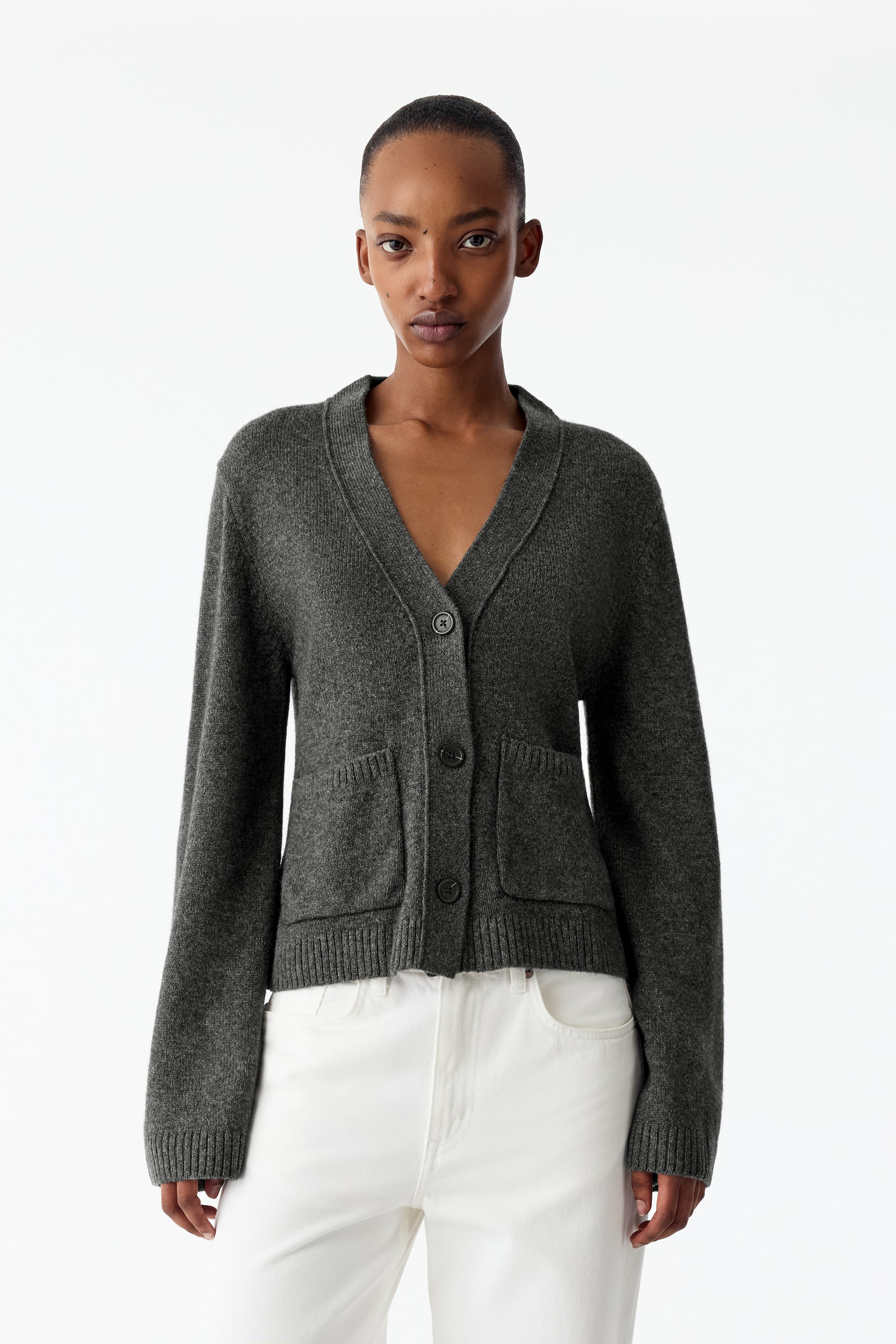 Visa större bild: En mörkgrå stickad cardigan med V-ringning, tre knappar framtill och två påsydda fickor fram bärs av modellen. Den slim fit-passformade cardigansmodellen är kombinerad med vita byxor.