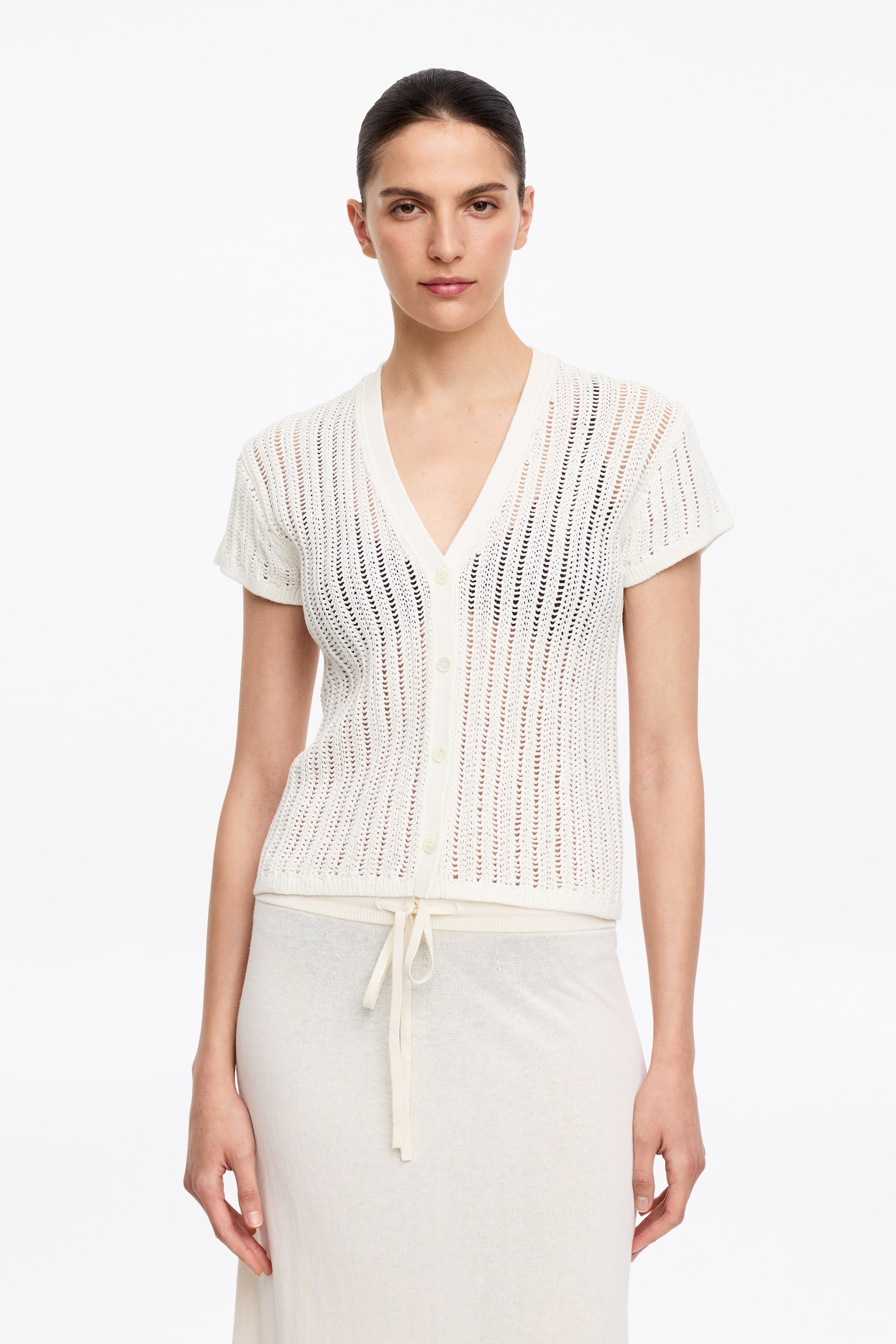 Agrandir l'image: Un cardigan blanc à manches courtes est présenté, avec un motif en maille ajourée et des œillets verticaux. Il est conçu avec un décolleté en V et une fermeture à boutons à l'avant, laissant entrevoir un haut foncé en dessous.
