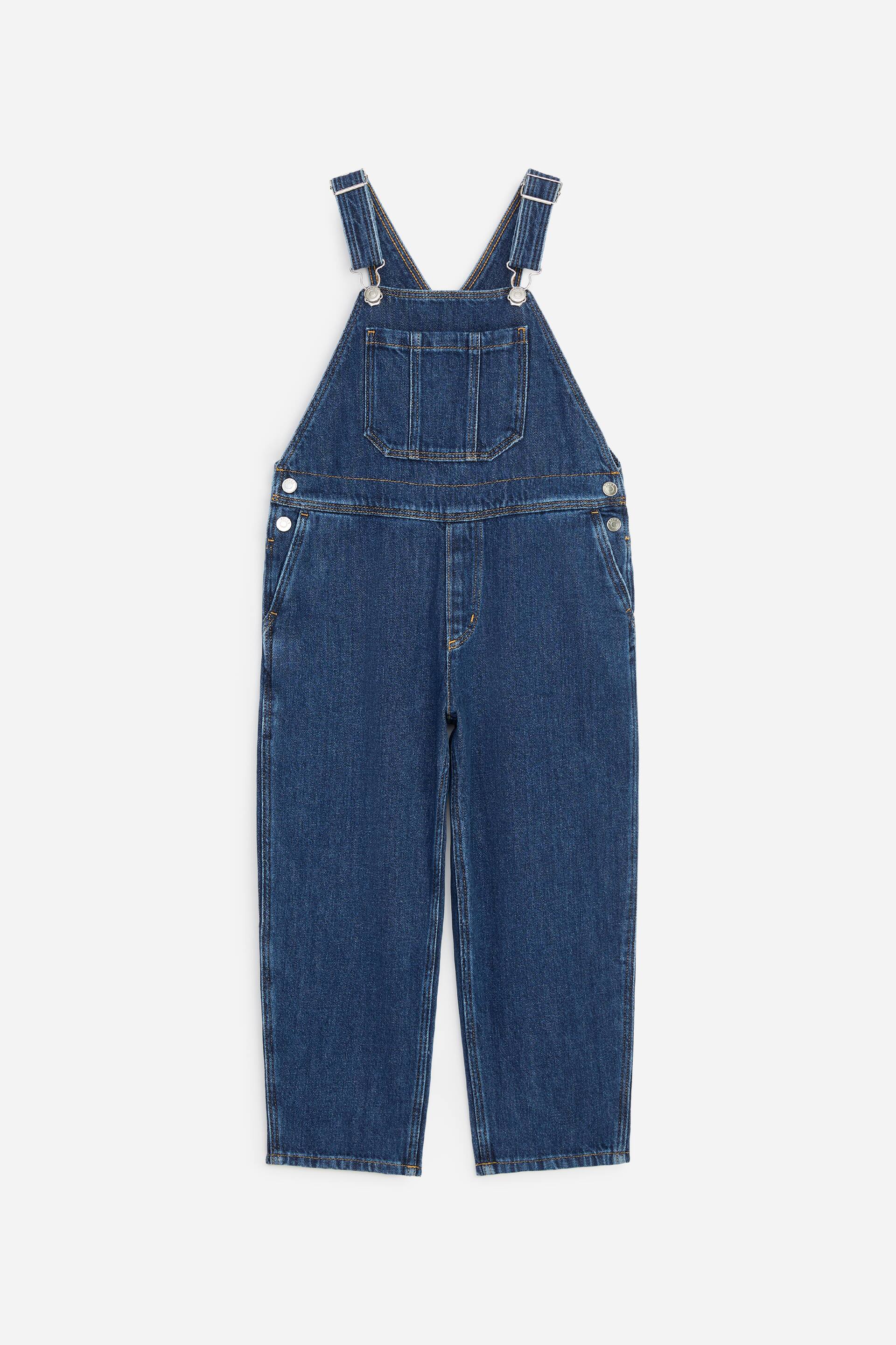 Agrandir l'image: Ce salopette en denim présente une silhouette à jambes barrel vue de face. Le modèle est doté de bretelles réglables avec boucles métalliques argentées, d’une poche plaquée centrale sur la poitrine et de poches latérales en biais.