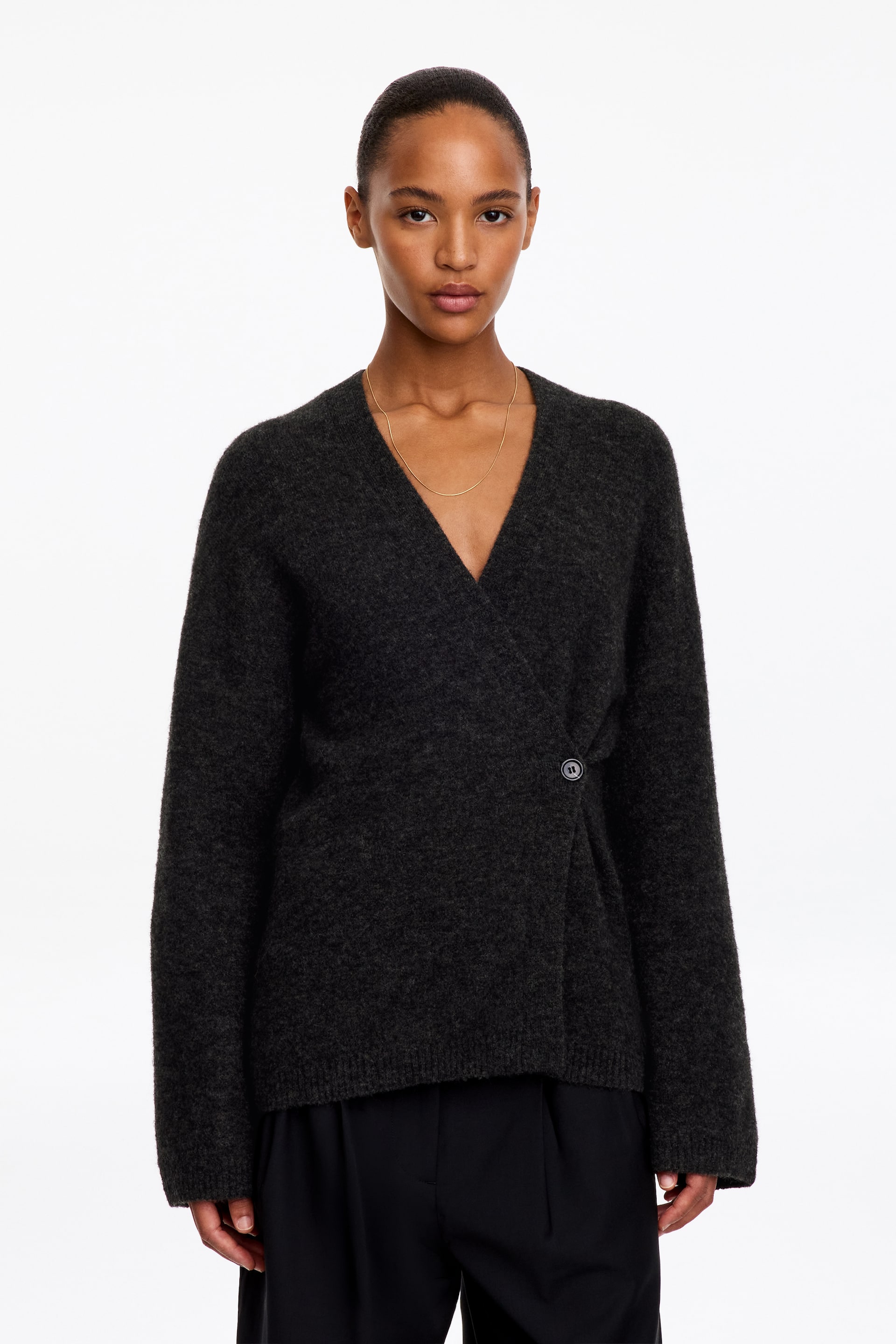 Agrandir l'image: Un cardigan cache-cœur à texture duveteuse gris foncé est porté par le mannequin, avec un profond col en V et une fermeture à un seul bouton sur le côté. Le cardigan est associé à un pantalon noir.