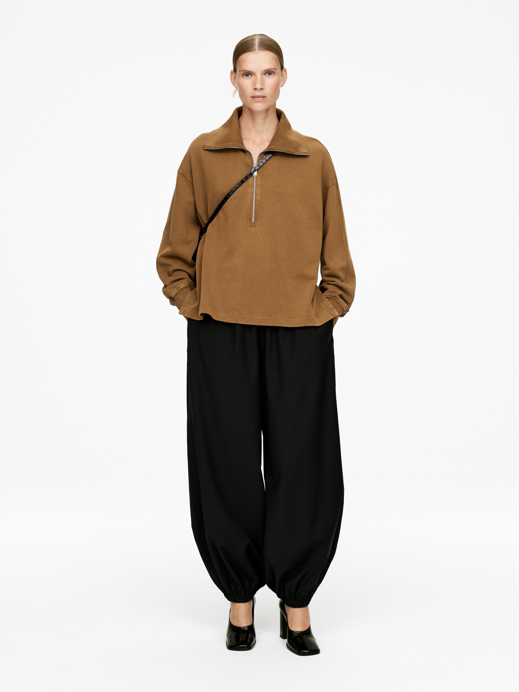 Grotere afbeelding bekijken: Een oversized camelbruine half-zip trui met een opstaande kraag en lange mouwen wordt gepresenteerd in een vooraanzicht. Het wordt gestyled met loszittende zwarte ballonbroeken, die bij de enkels samenkomen, en zwarte lakleren schoenen met hoge hakken, die de ontspannen silhouet van het ensemble laten zien.