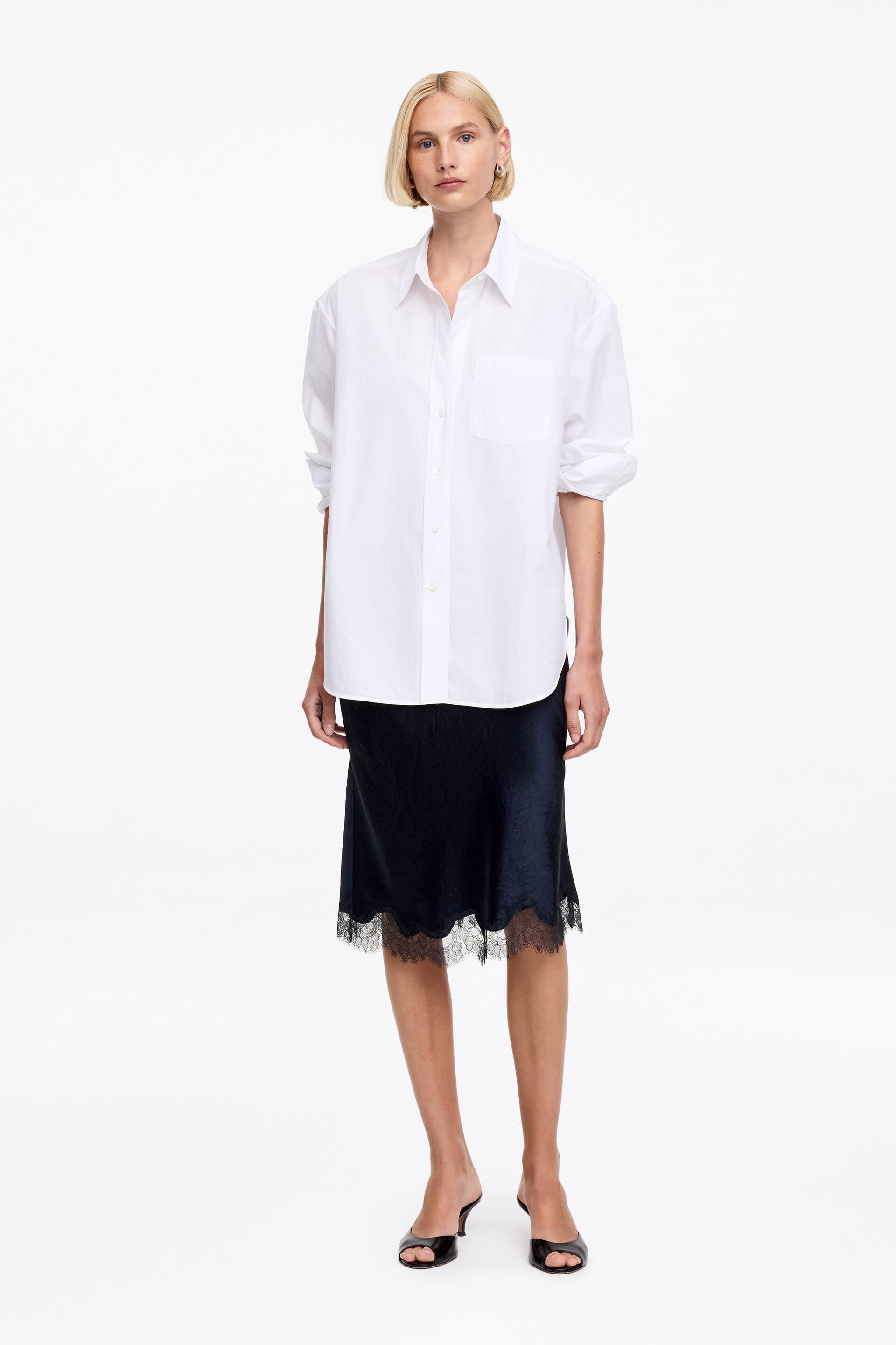 Visualizza immagine più grande: Una camicia bianca oversize con bottoni, caratterizzata da un colletto a punta, maniche lunghe arrotolate e una tasca a toppa sul petto, viene presentata abbinata a una gonna sottoveste nera in raso di lunghezza midi, che mostra un delicato bordo in pizzo nero sull'orlo, con entrambi i capi rivolti verso il fronte.
