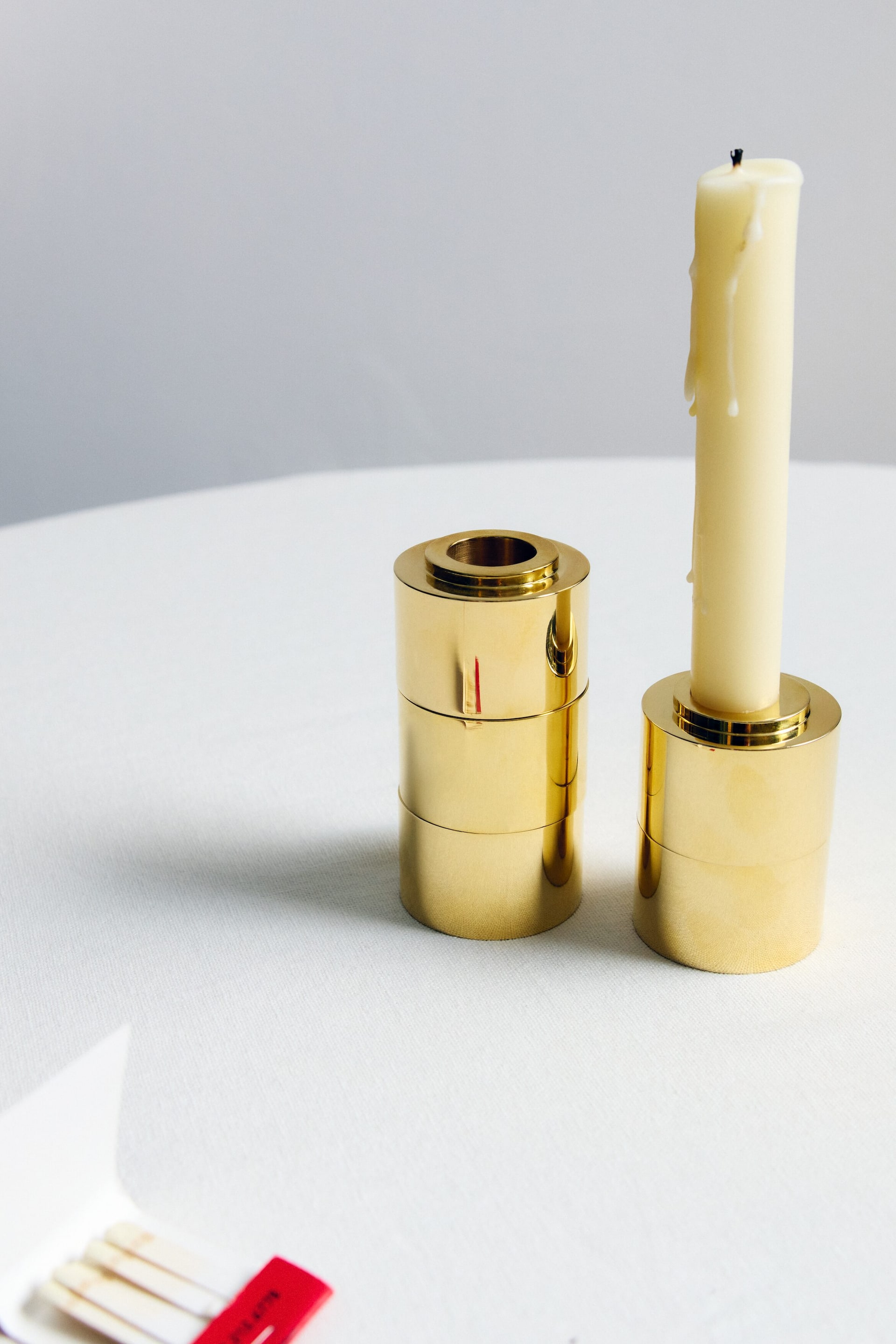 View larger image: Two cylindrical, polished brass candleholders are standing upright. Each holder features a horizontal seam, suggesting a stacked design. One holder displays a cream-coloured taper candle, showing visible wax drips and a burnt wick, while the other holder is empty, revealing its central opening.