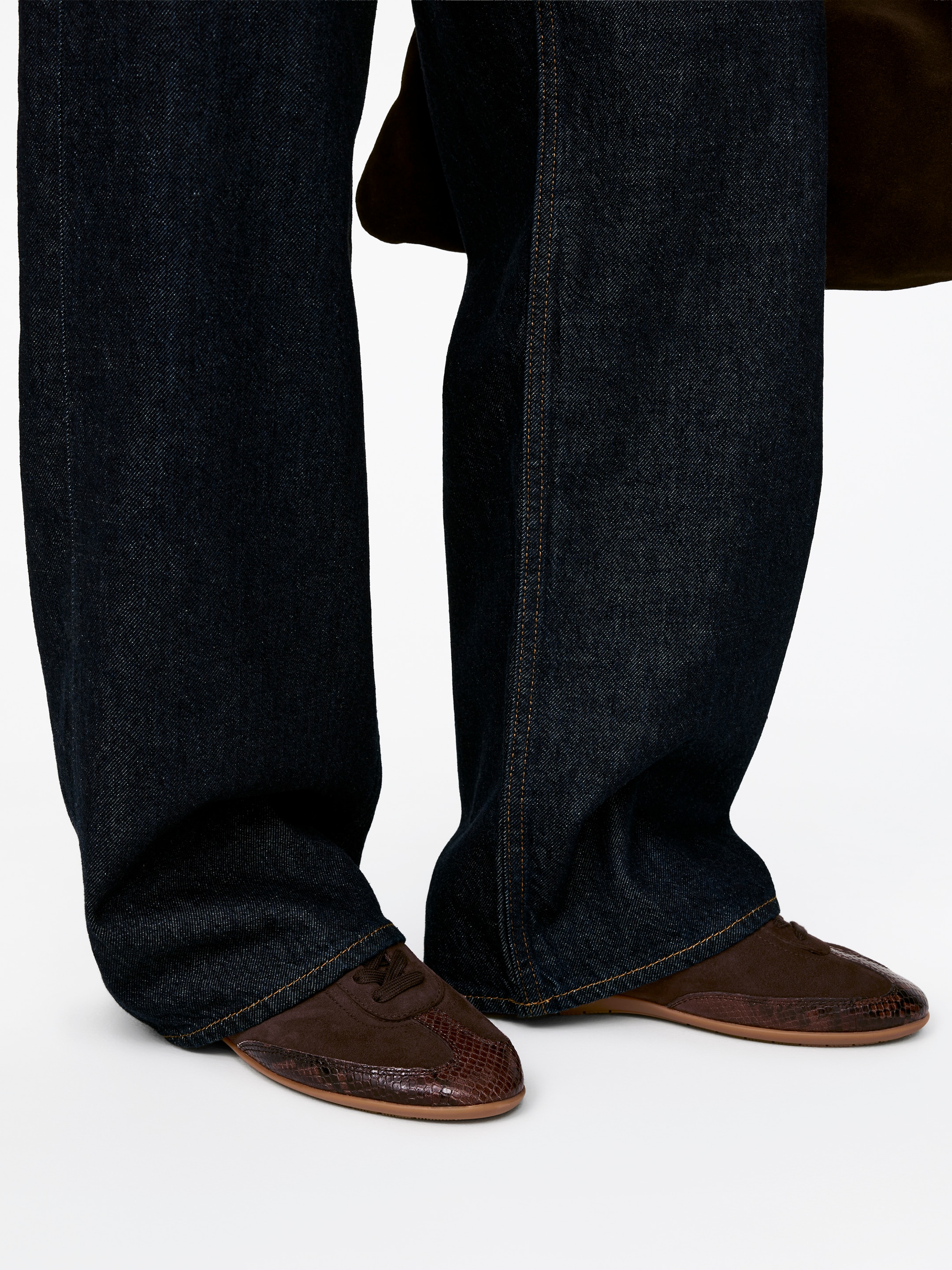 View larger image: Trainers, crafted from brown suede with a darker, textured toe cap, are being worn by the model. These streamlined trainers feature lace-up fastening, and dark blue wide-leg jeans are covering their upper portion.