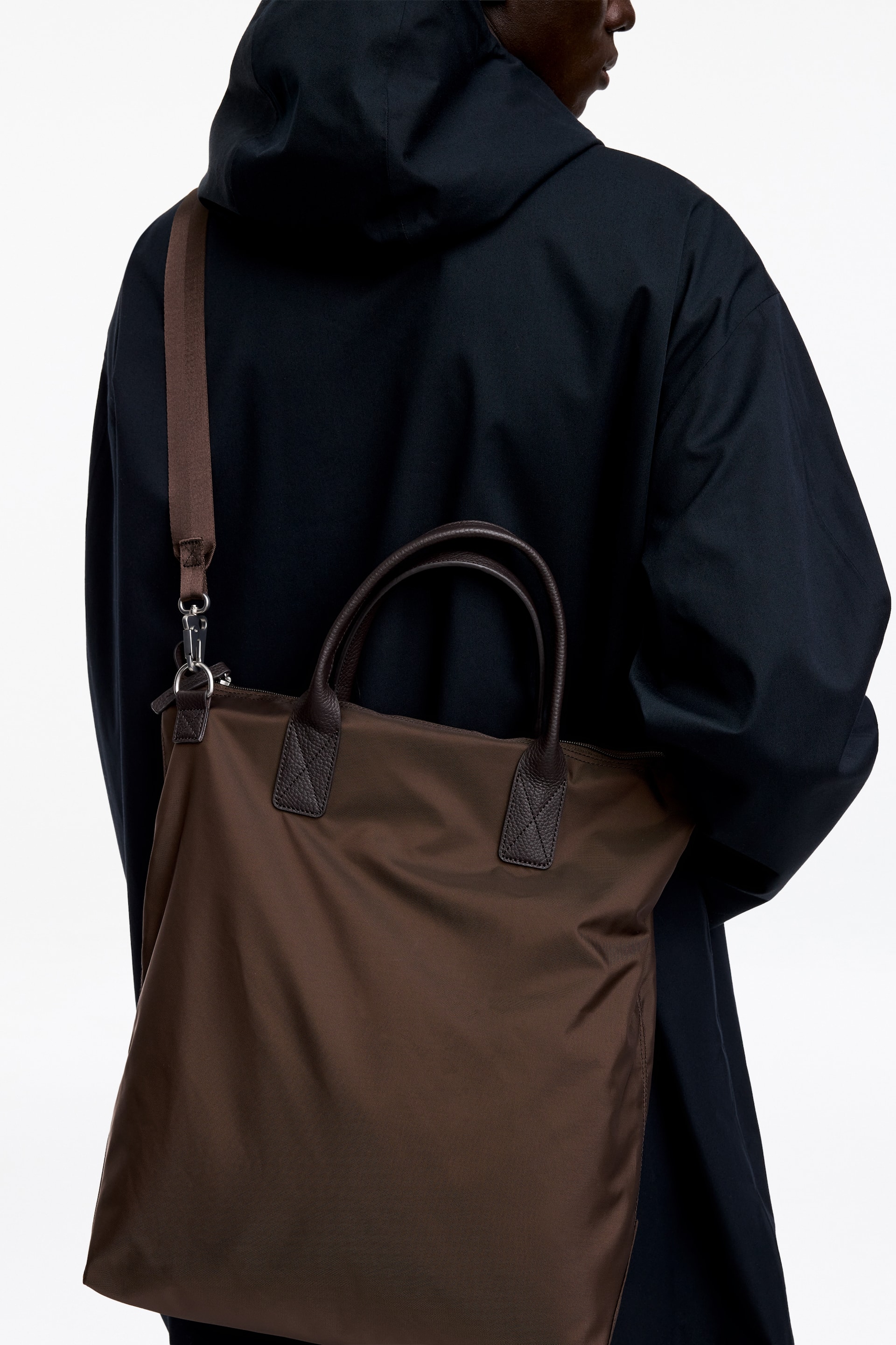 View larger image: A brown nylon tote bag featuring dark brown leather top handles and a matching fabric shoulder strap is being carried by the model over their shoulder. The bag rests against a dark hooded jacket worn by the model.