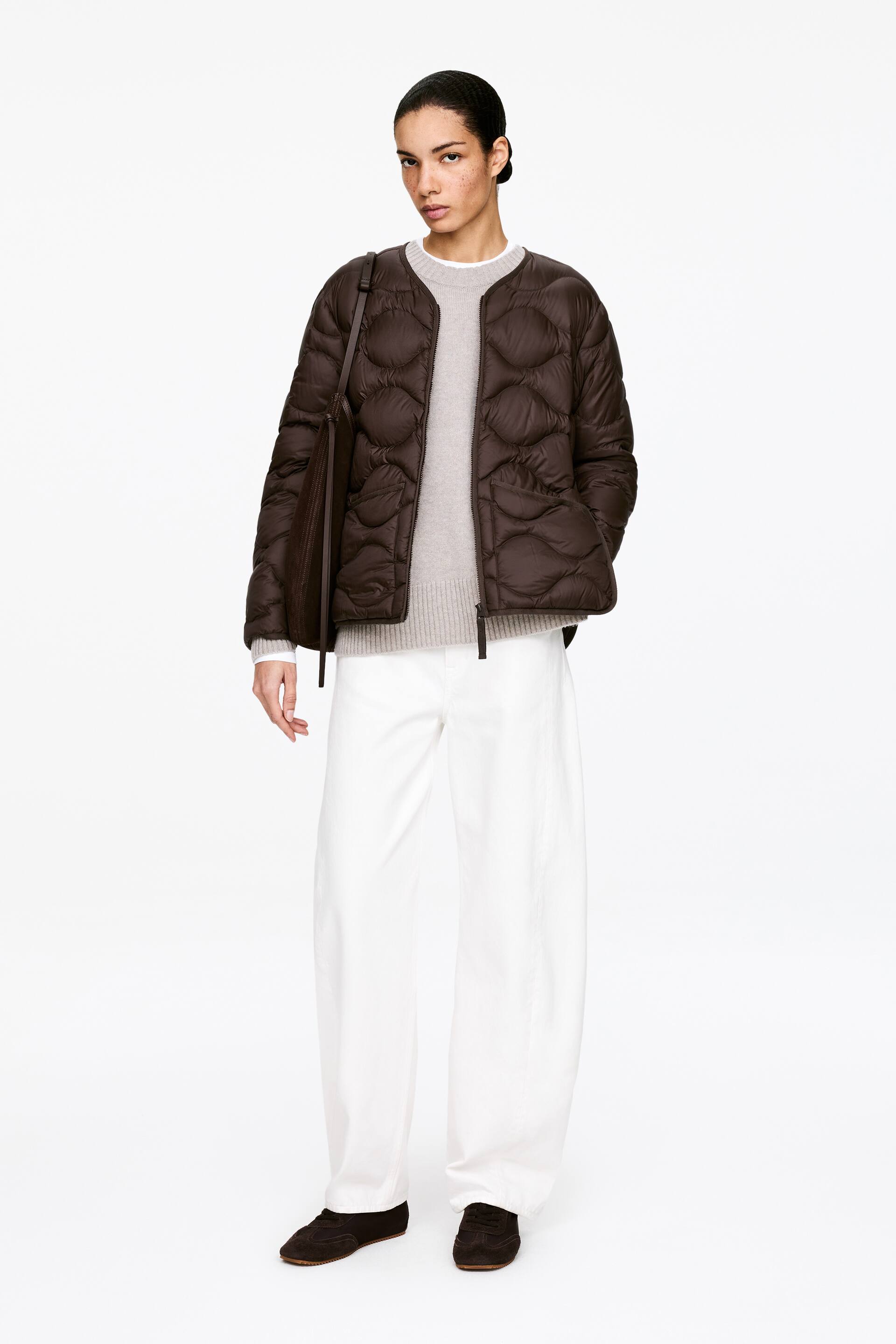 Größeres Bild anzeigen: Eine braune, gesteppte Jacke wird präsentiert, die frontal und leicht nach rechts gewinkelt ist. Sie verfügt über einen kragenlosen, runden Ausschnitt und einen durchgehenden Reißverschluss vorne. Sie ist mit einem organischen, wellenförmigen Steppmuster und zwei aufgesetzten Taschen vorne gestaltet und wird über einem hellgrauen Rundhalspullover getragen.