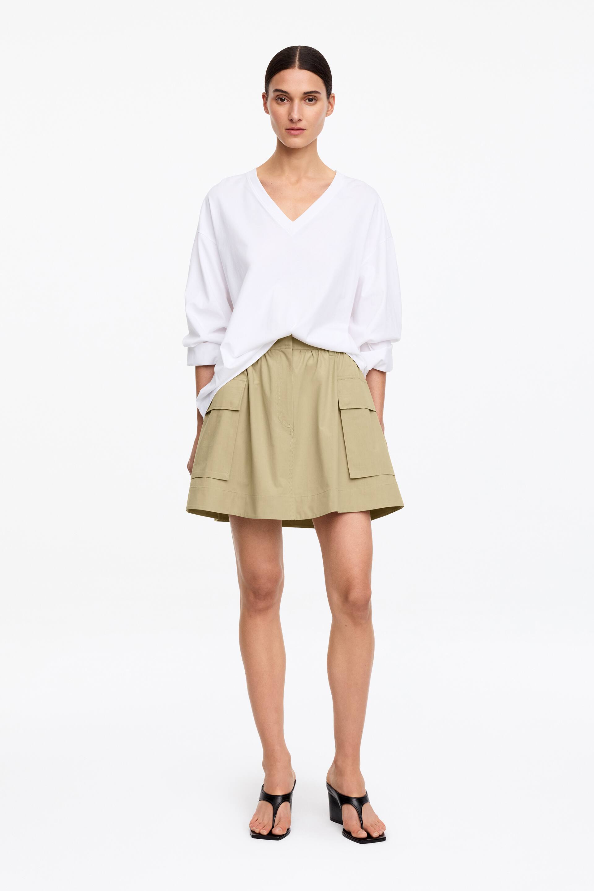 Visa större bild: En ensemble innehåller en lös vit V-ringad topp med trekvartsärmar, avslappnat instoppad i en khaki minikjol. Kjolen är designad med en A-linje siluett, en elastisk midja och två stora ficklock på framsidan, och kompletteras med svarta thong kilklackssandaler.