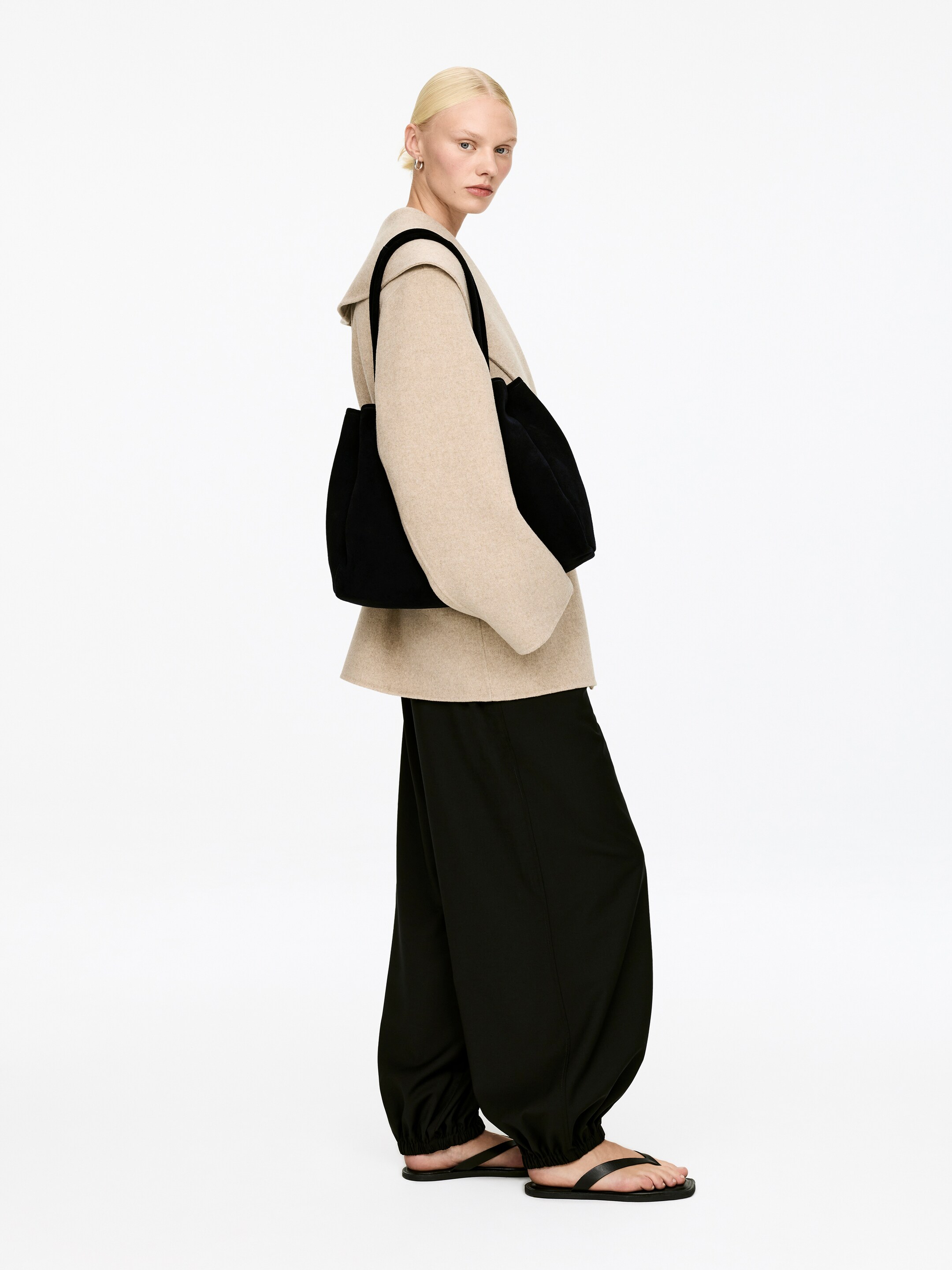 Agrandir l'image: Un sac cabas noir est porté sur l'épaule droite d'une personne, avec son corps principal orienté vers l'avant. Il présente un design doux et non structuré avec des fronces verticales sur les côtés, créant une apparence arrondie et pleine.