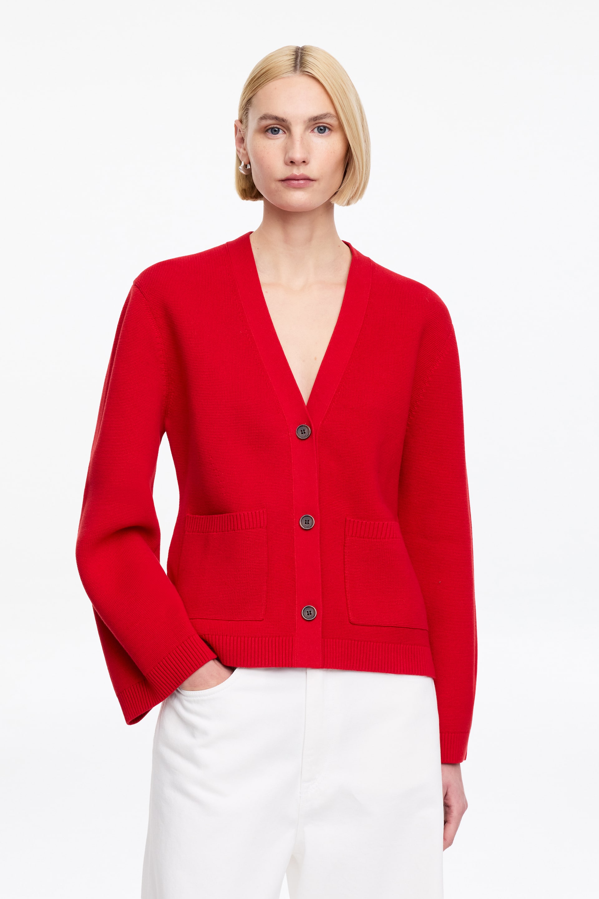 Vezi imaginea mai mare: Un cardigan roșu este orientat cu fața înainte, având un decolteu în V și trei nasturi rotunzi, închiși la culoare, pe partea din față. Prezintă două buzunare aplicate aproape de tiv, mâneci lungi cu manșete nervurate și un tiv nervurat.