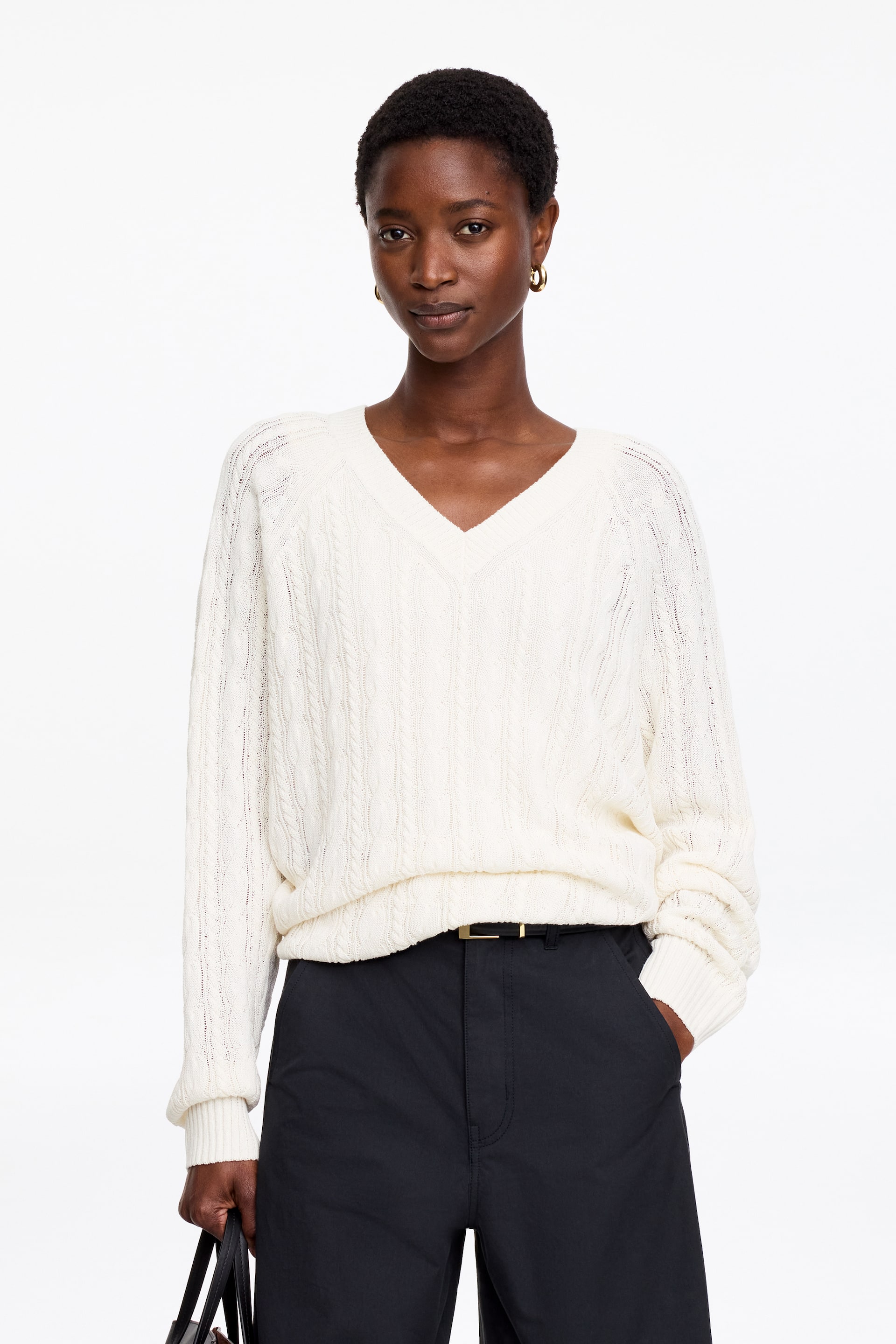 Ver imagen más grande: Un jersey de punto trenzado color crema con cuello en V, que presenta acabados acanalados, está siendo llevado por la modelo. El jersey de corte holgado se combina con pantalones oscuros, y la modelo lleva un bolso oscuro.