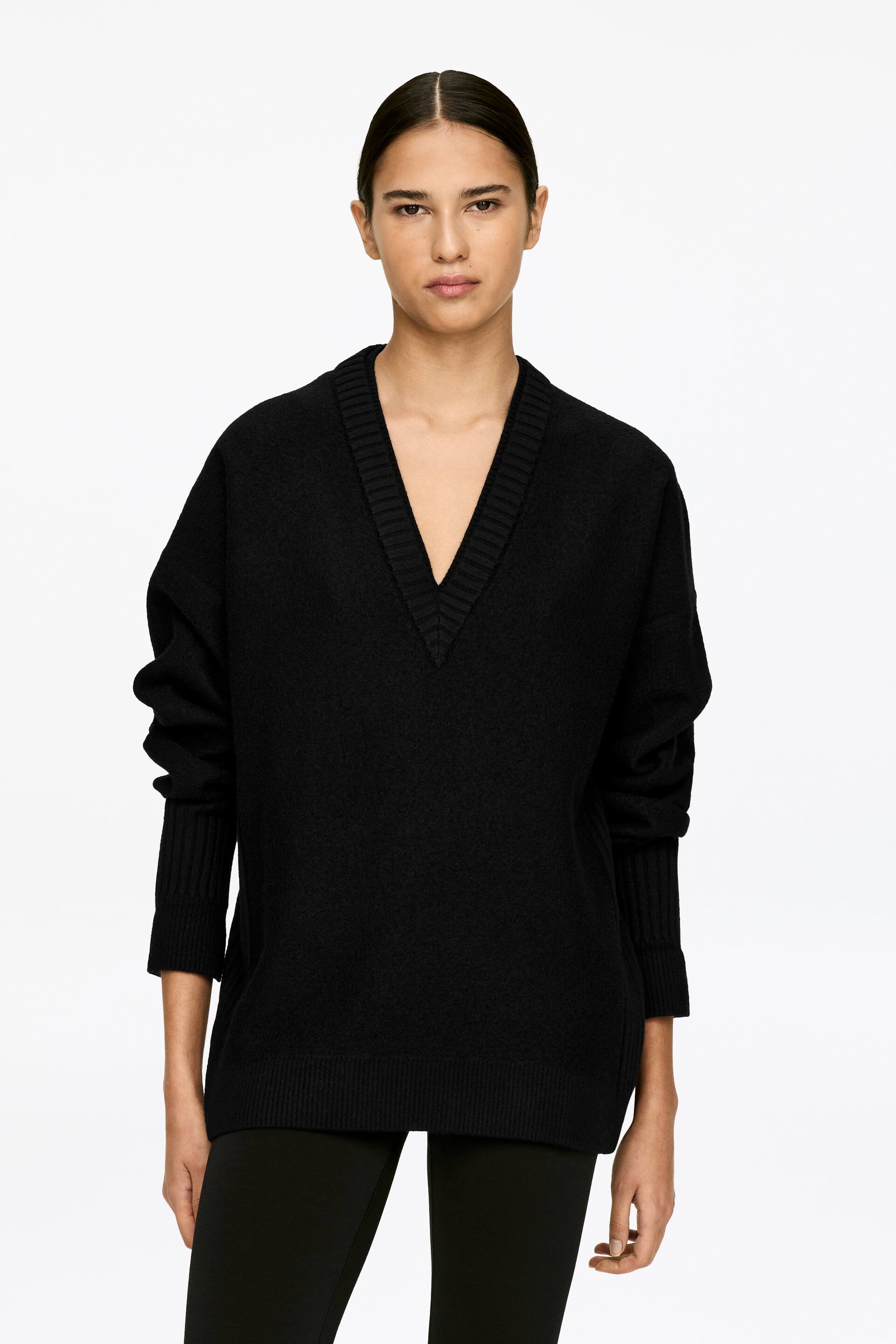 Größeres Bild ansehen: Ein schwarzer Pullover aus gekochter Wolle wird vom Model getragen. Er ist mit einem doppellagigen V-Ausschnitt, umgeschlagenen Bündchen und einem gerippten Saum versehen.