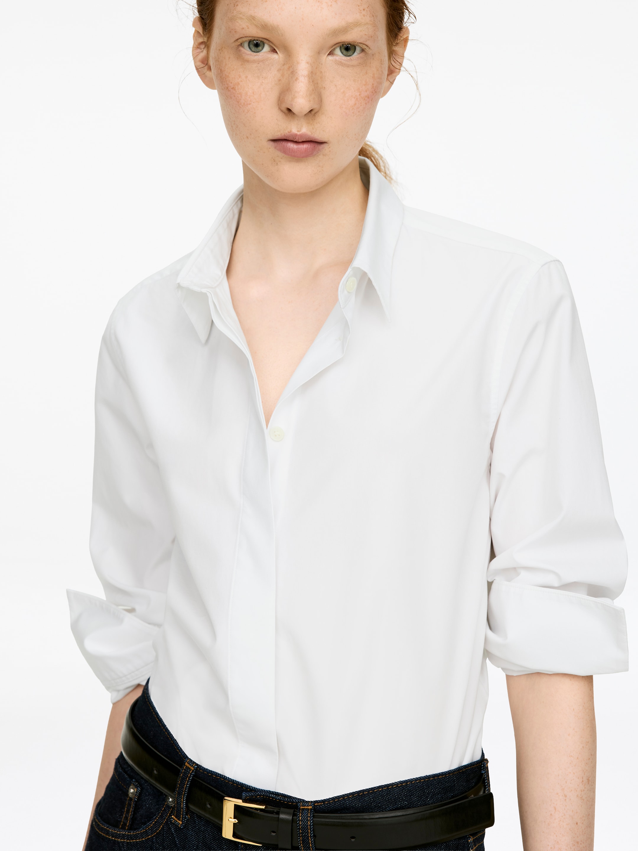 Visualizza immagine più grande: Una camicia classica bianca di cotone con una vestibilità regolare e un'allacciatura frontale con bottoni nascosti viene indossata dalla modella, il colletto a punta aperto e le maniche lunghe arrotolate ai polsini. La camicia è infilata in pantaloni scuri e fissata con una cintura nera.