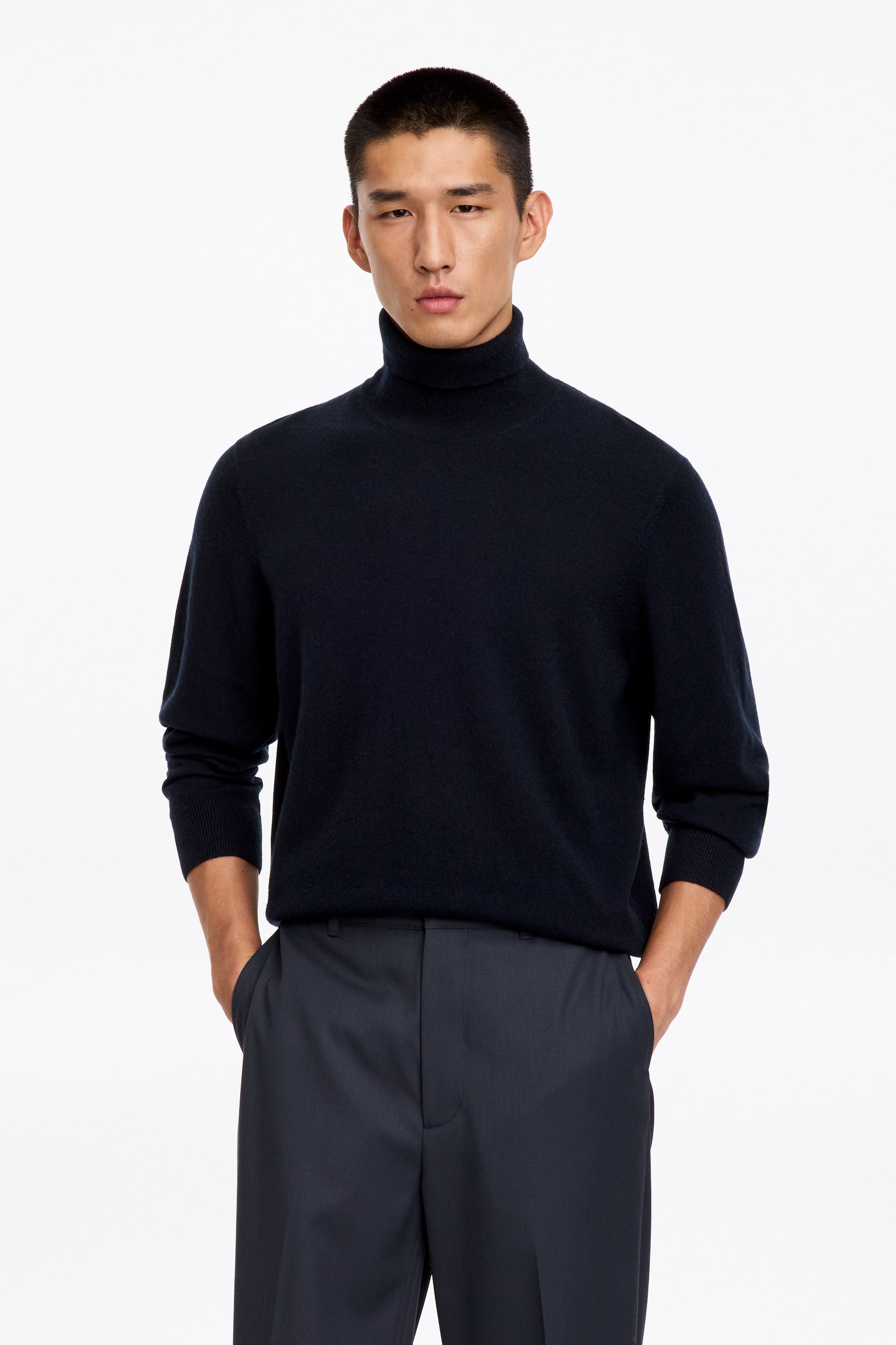 Agrandir l'image: Le mannequin porte un pull à col roulé bleu marine, debout avec les mains dans les poches d’un pantalon gris foncé. Ce pull en maille unie présente un col montant côtelé et des poignets côtelés remontés sur les avant-bras.