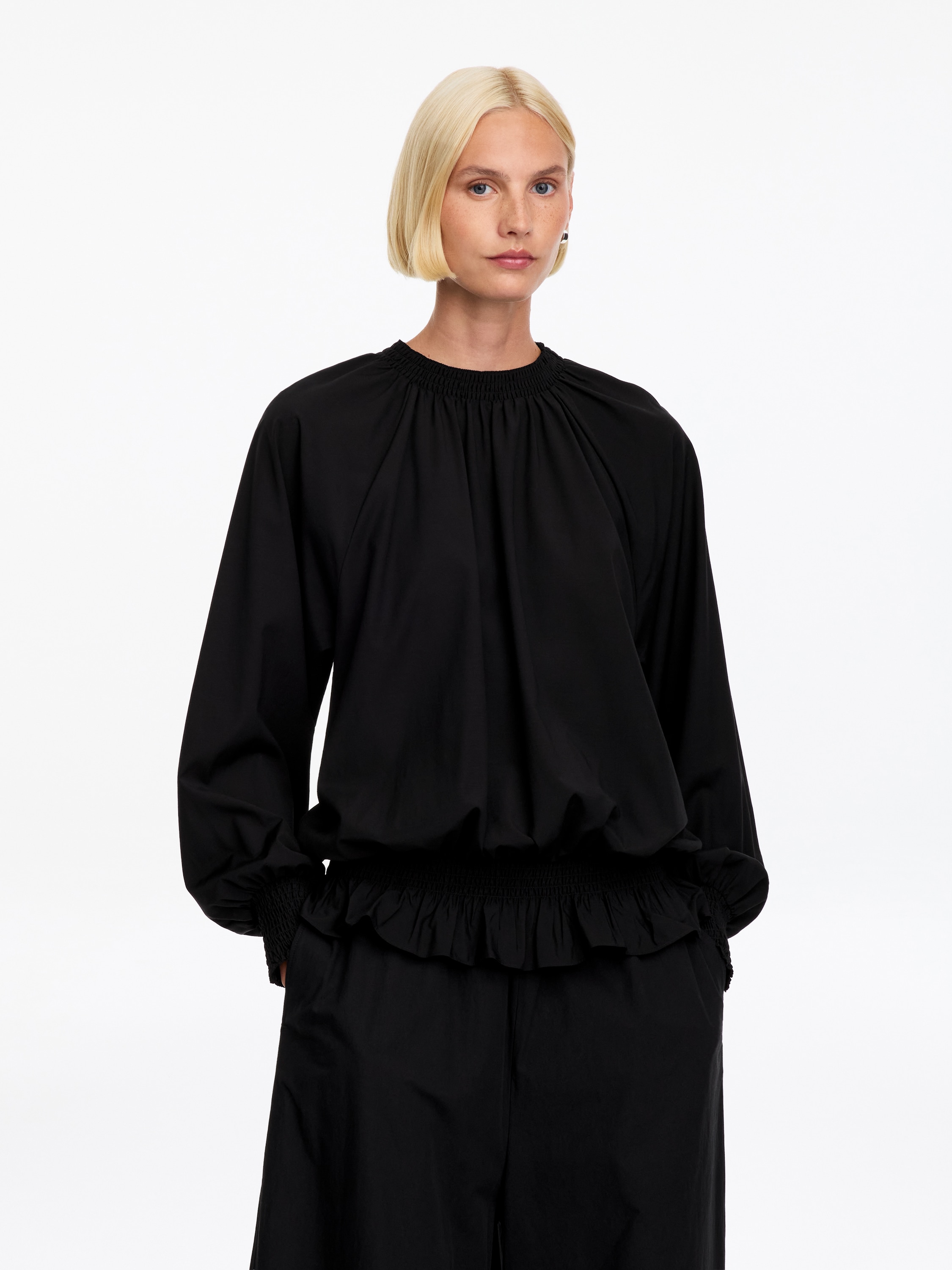 En svart bluse med lange ermer er vist forfra, med hÃ¸y, rynket hals og et bredt, rynket midjebÃ¥nd som skaper en samlet blouson-silhuett med volangkant. Blusen er designet med voluminÃ¸se ermer som avsluttes med matchende brede, rynkede mansjetter.
