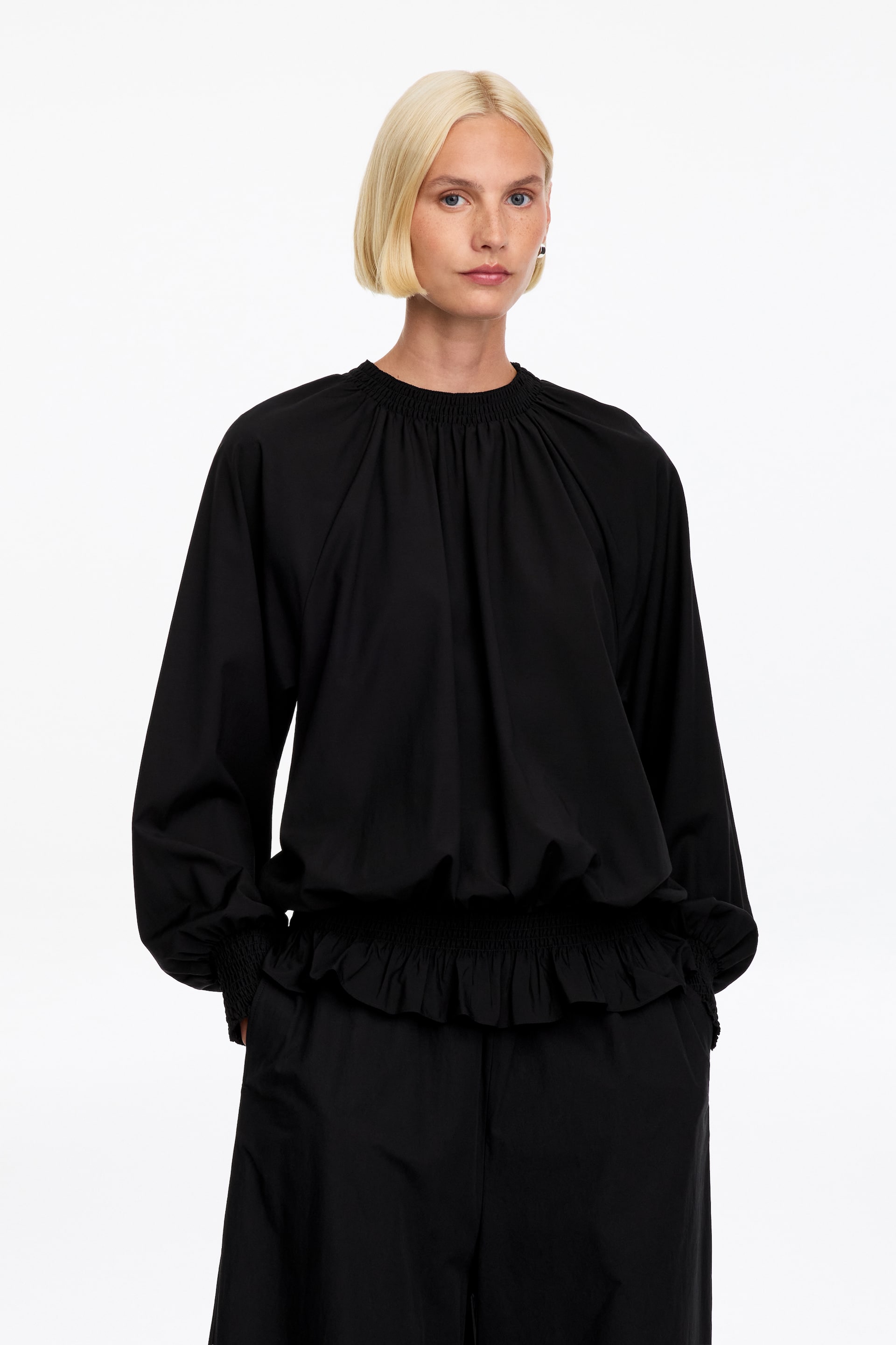 Visualizza immagine più grande: La modella indossa una blusa nera a maniche lunghe, caratterizzata da un collo alto arricciato, maniche voluminose con polsini arricciati e un orlo elasticizzato arricciato con bordo volant. La blusa è abbinata a pantaloni neri coordinati.