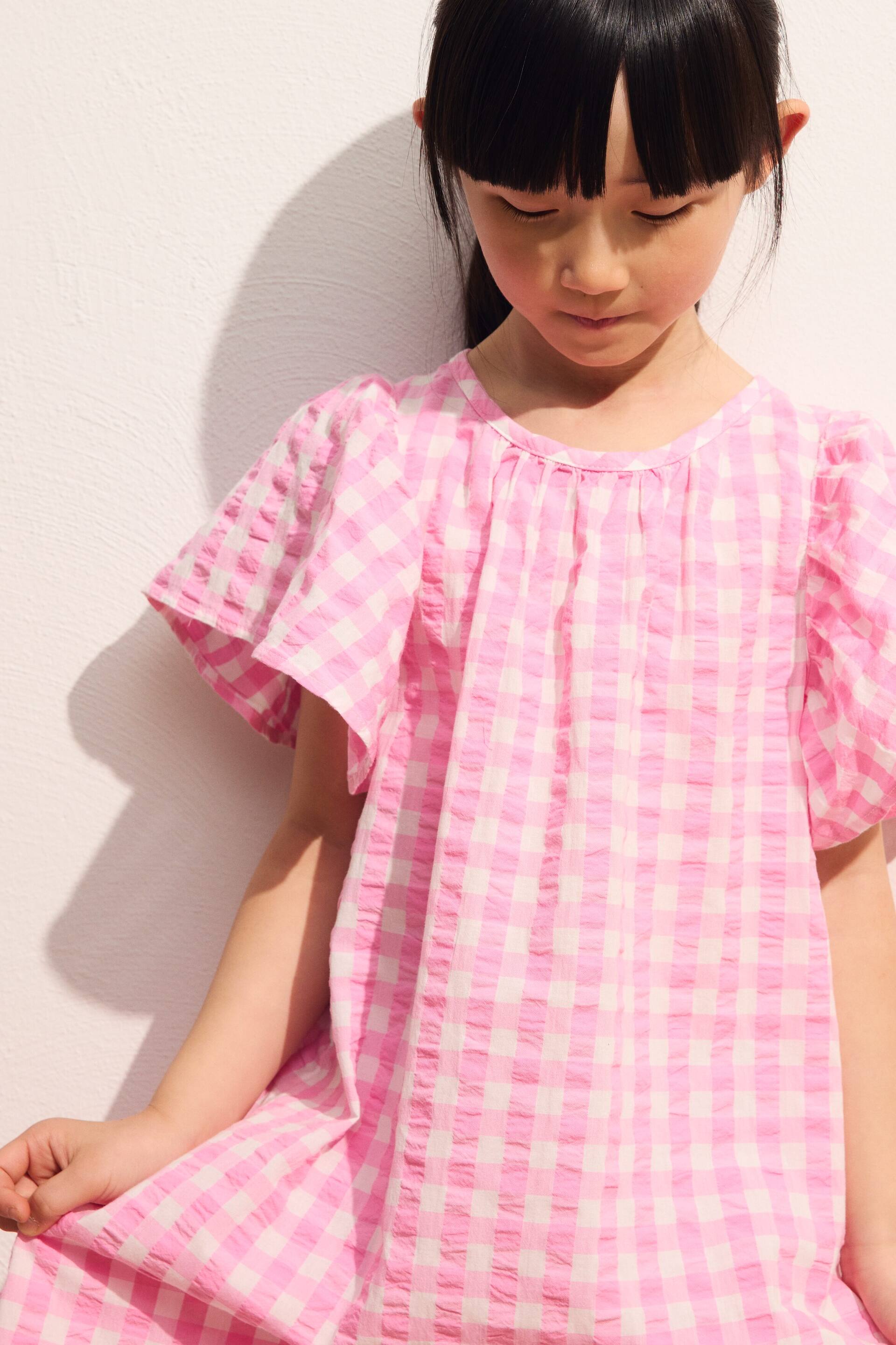 Größeres Bild ansehen: Ein rosa-weiß kariertes Kleid mit Vichy-Muster präsentiert einen rund gerafften Halsausschnitt und kurze Flatterärmel. Das Kleid zeigt eine geknitterte Textur und einen lockeren Schnitt.