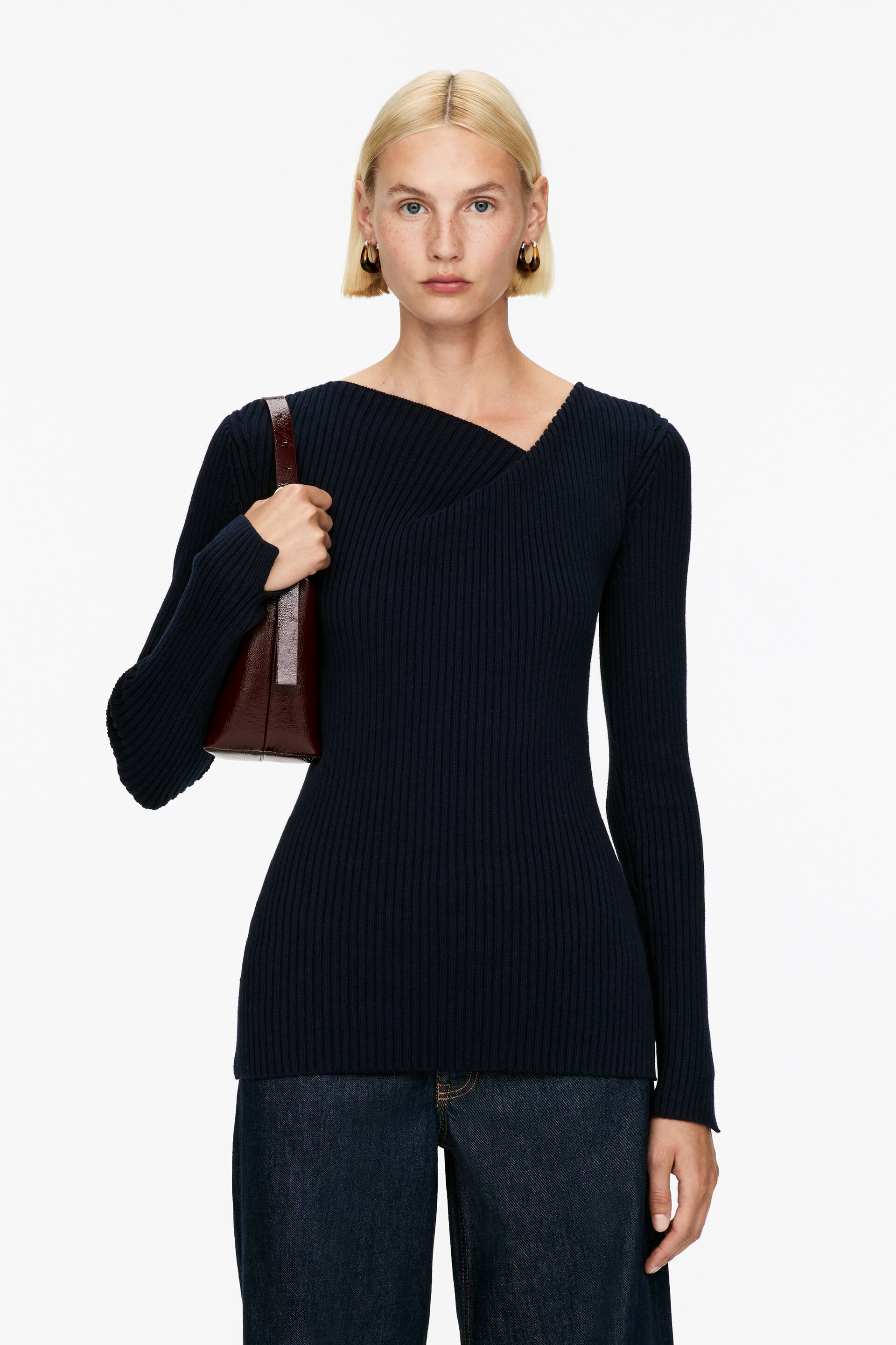 Vis større bilde: En mørkeblå, langermet ribbestrikket topp presenteres forfra. Toppen har en asymmetrisk V-hals med en kryssdetalj, og den viser en smal passform som strekker seg til hoften.