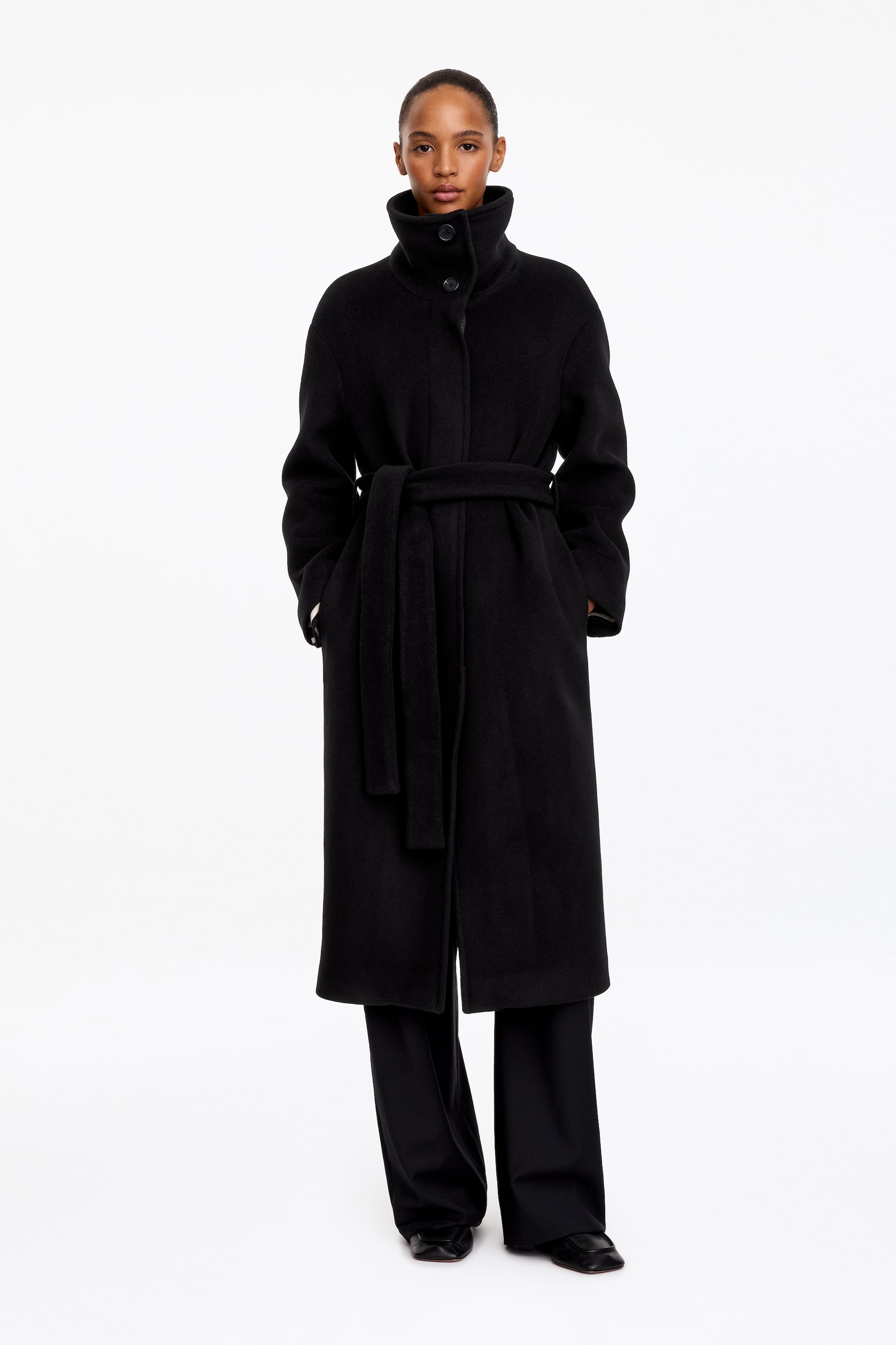 View larger image: A long black wool-blend coat is worn by the model, featuring a high stand collar and a matching fabric belt tied at the waist. The model stands with hands placed in the coat’s side pockets, styling it with wide-leg black trousers.