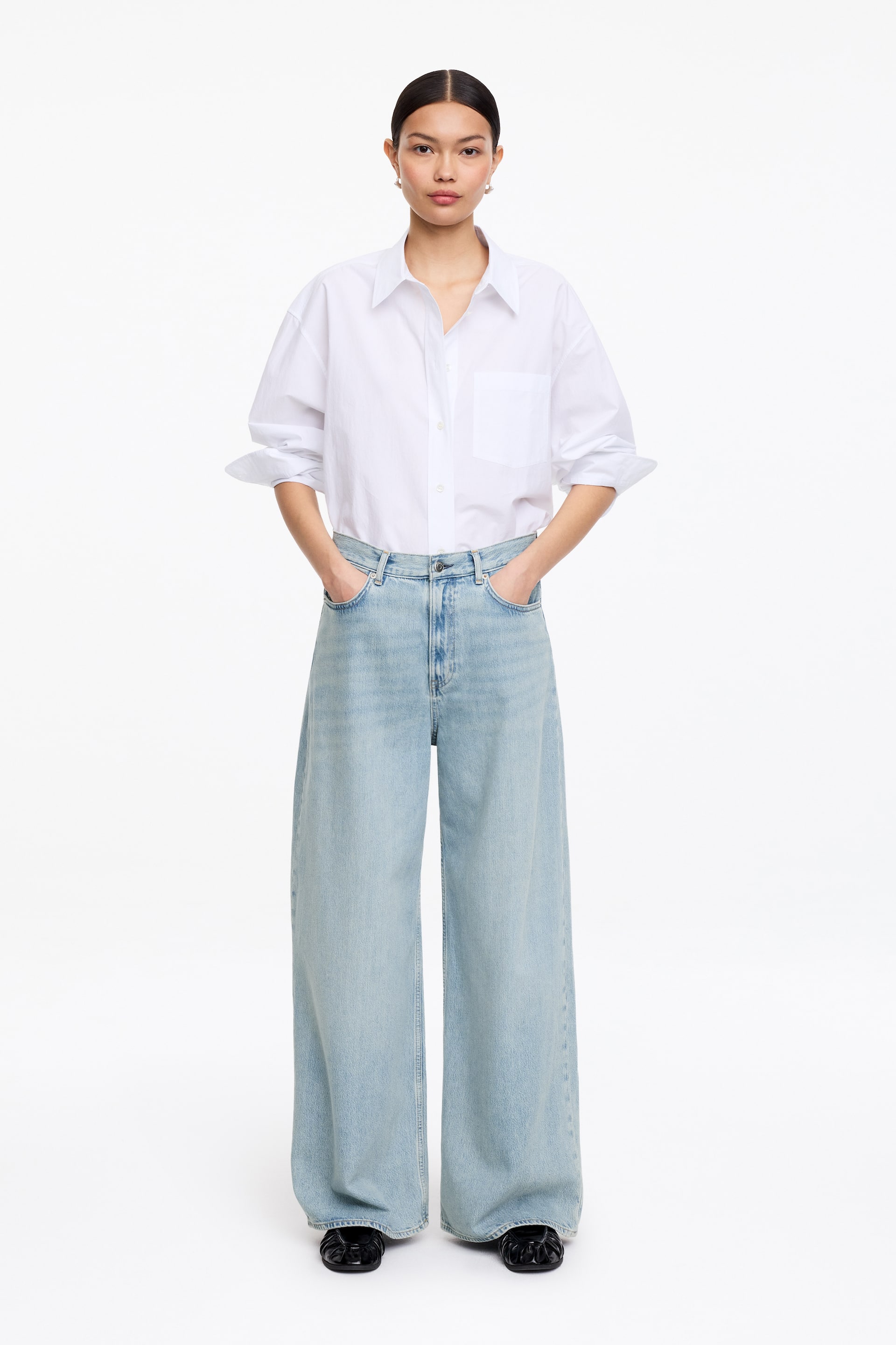 Visa större bild: En vit skjorta med knappar har en spetsig krage, en bröstficka och uppvikta ärmar, och visas instoppad i ljusa, högmidjade jeans med vida ben. Jeansen har en lös passform som sträcker sig till full längd och vilar över svarta skor.