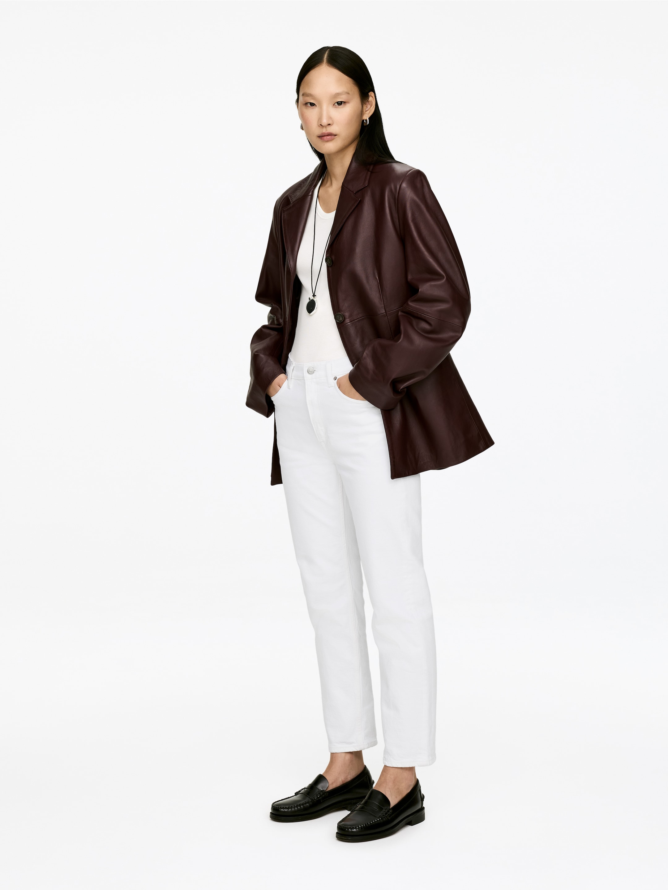 Agrandir l'image: Le modèle porte un jean en denim blanc coupé, avec une taille mi-haute, une coupe ajustée et des jambes fuselées qui s'arrêtent au-dessus de la cheville. Le modèle est debout, les mains dans les poches du jean, les stylisant avec un haut blanc, une veste marron foncé et des mocassins noirs.